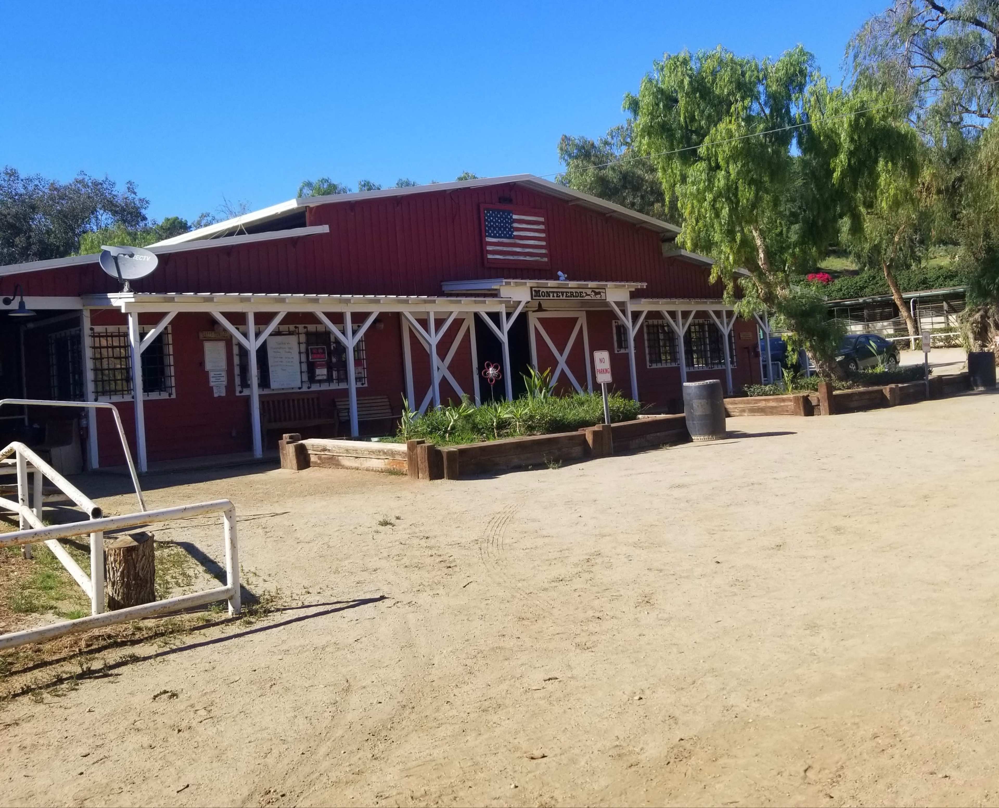 A red barn-like building with an American flag, surrounded by a dirt area and green trees.