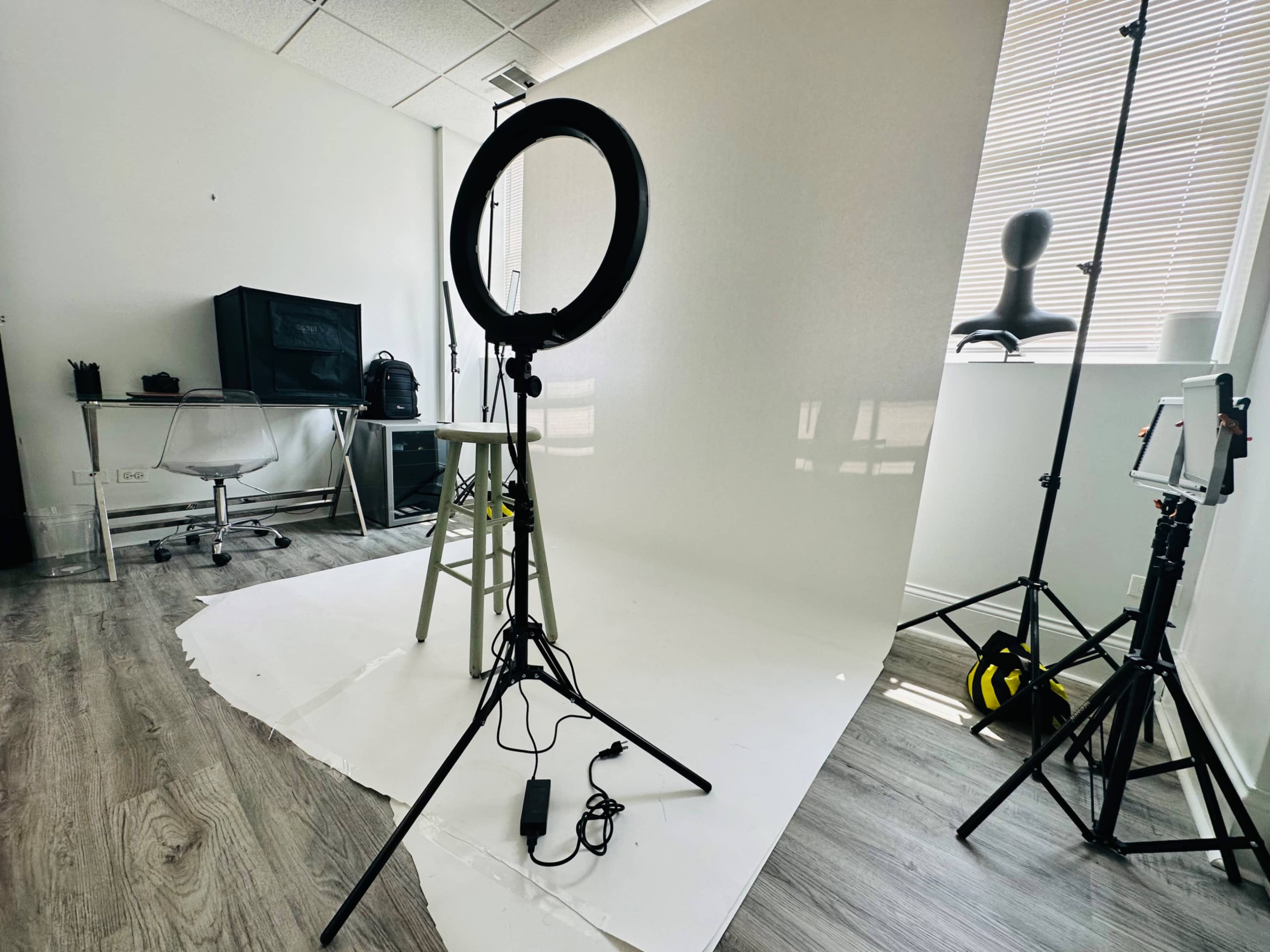 The image shows a photography studio setup featuring a ring light, a stool, and a white backdrop, with additional equipment and furniture in the background.