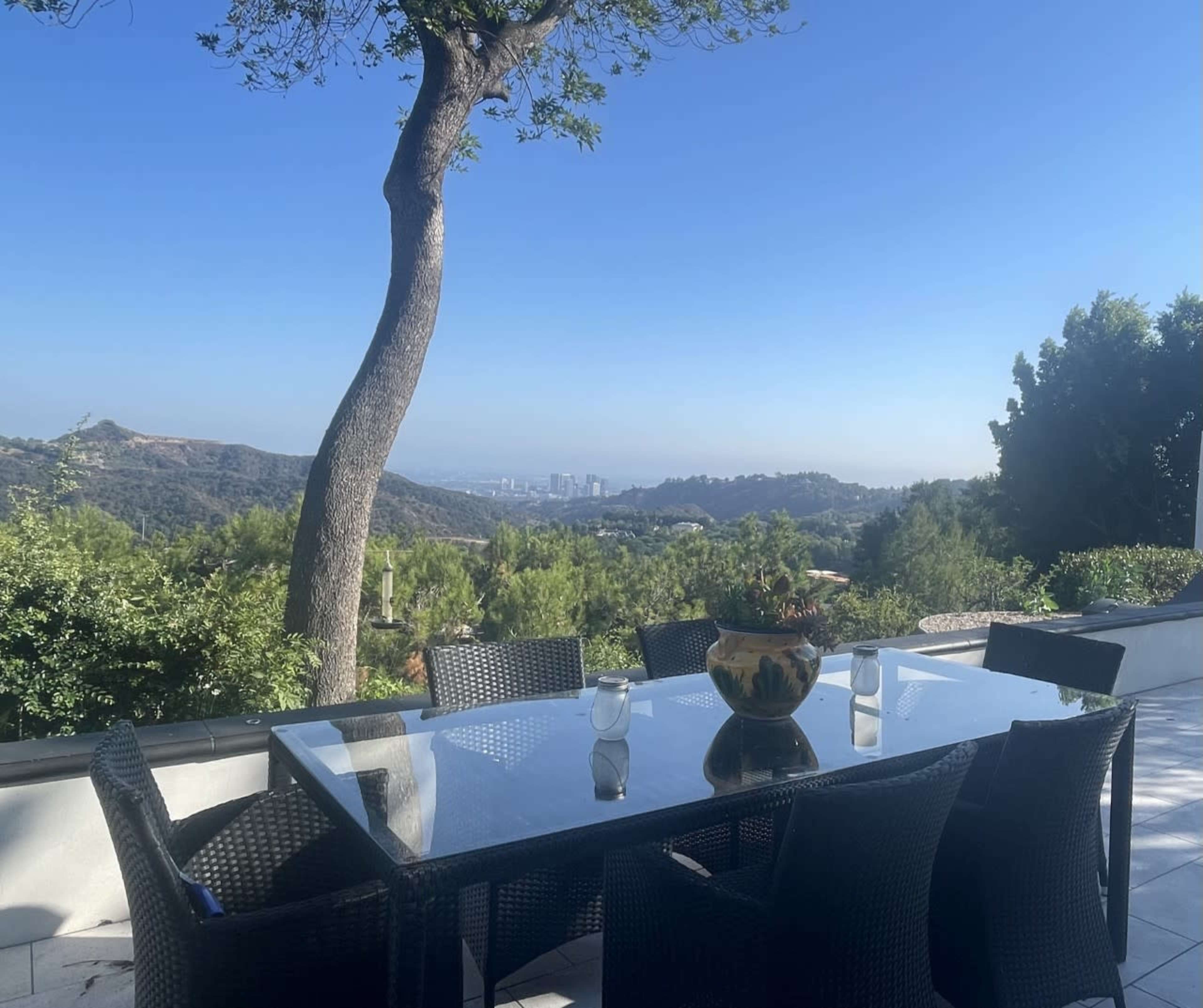 A glass-top dining table surrounded by black wicker chairs is set on a patio overlooking a scenic view of hills and a distant city skyline.