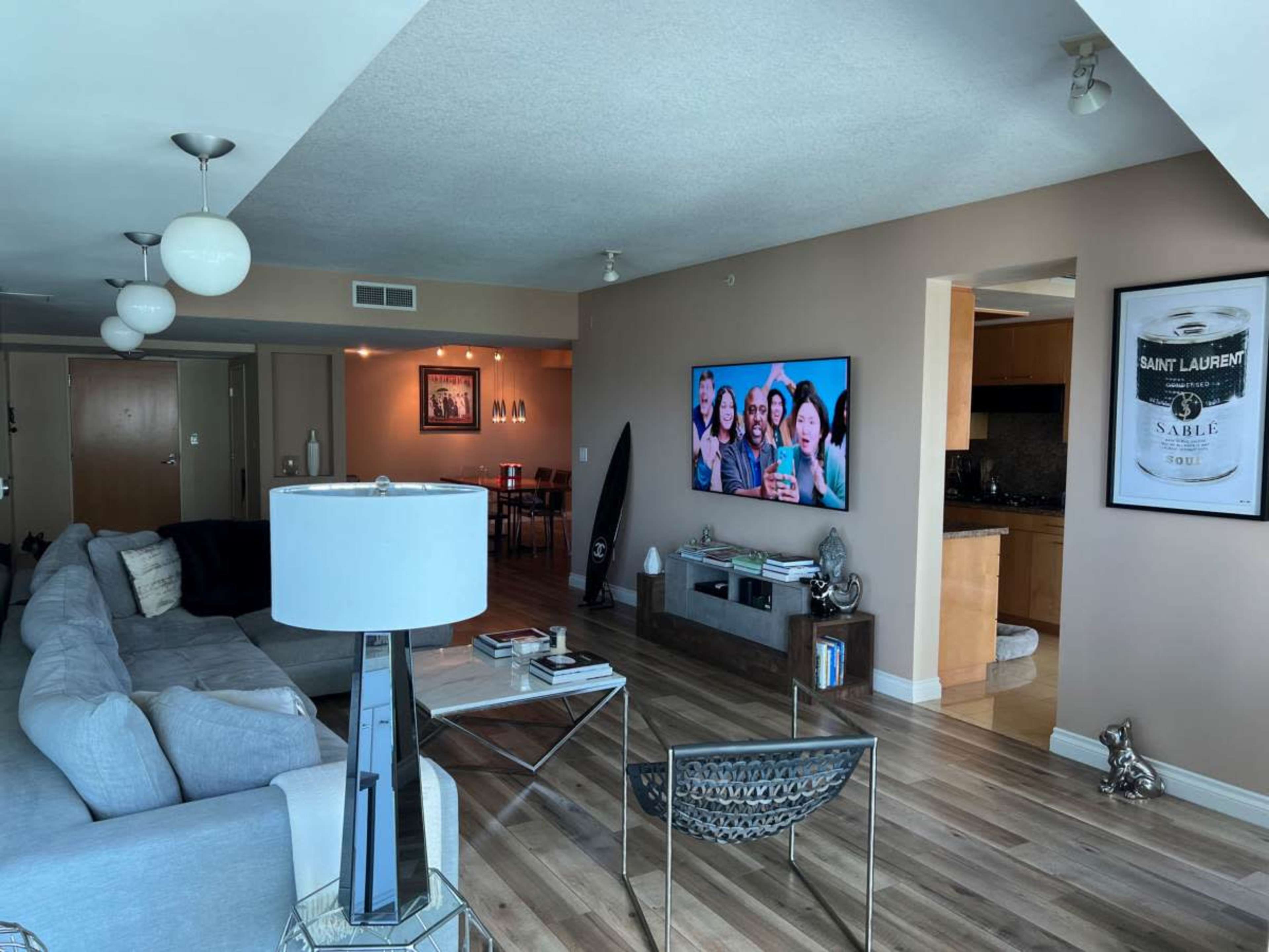 A modern living room with a gray sectional sofa, a TV mounted on the wall displaying a group photo, and a dining area visible in the background.