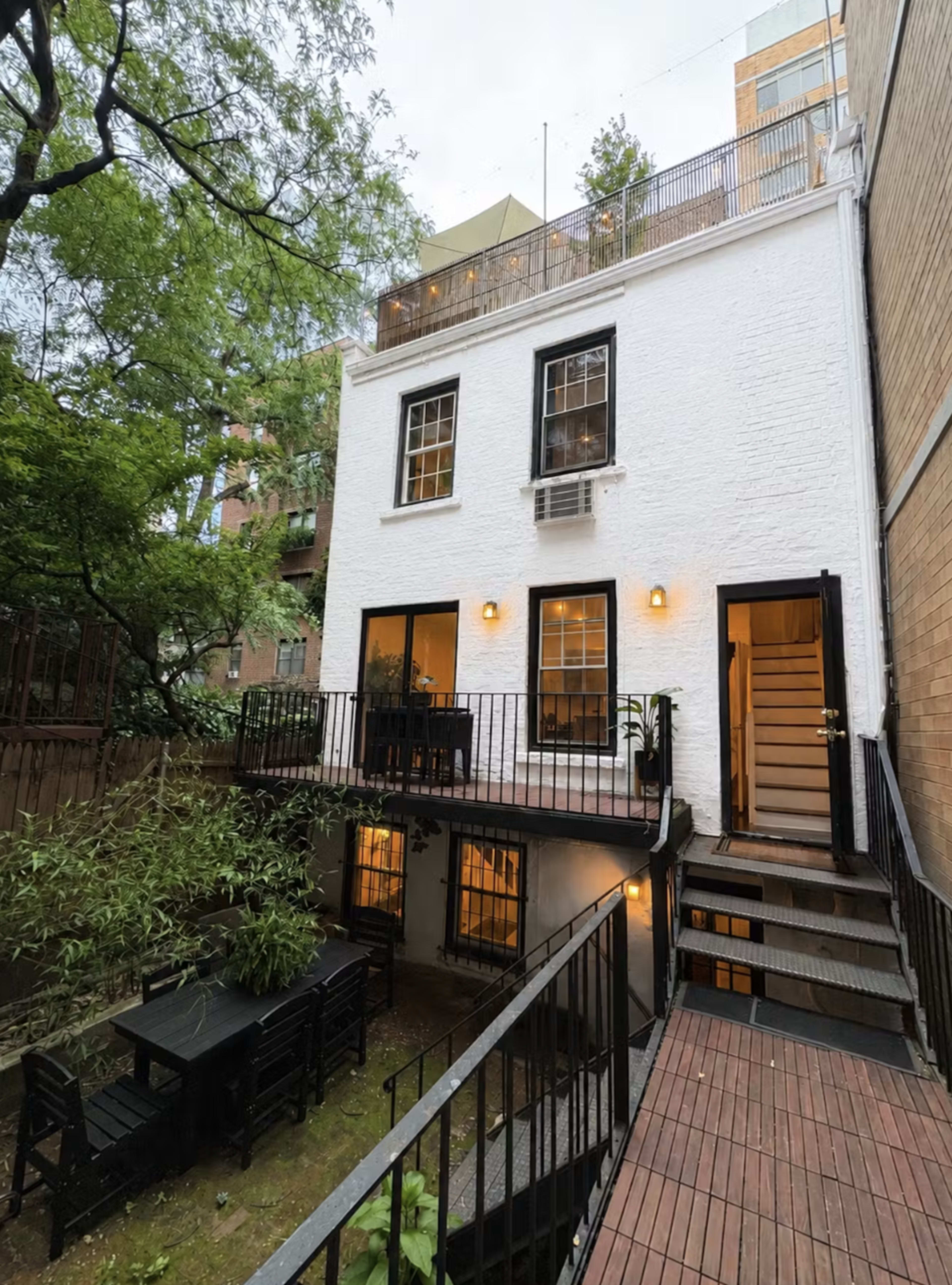 The image shows a two-story white residential building with black window frames and a wooden stairway leading to a small patio area surrounded by greenery.