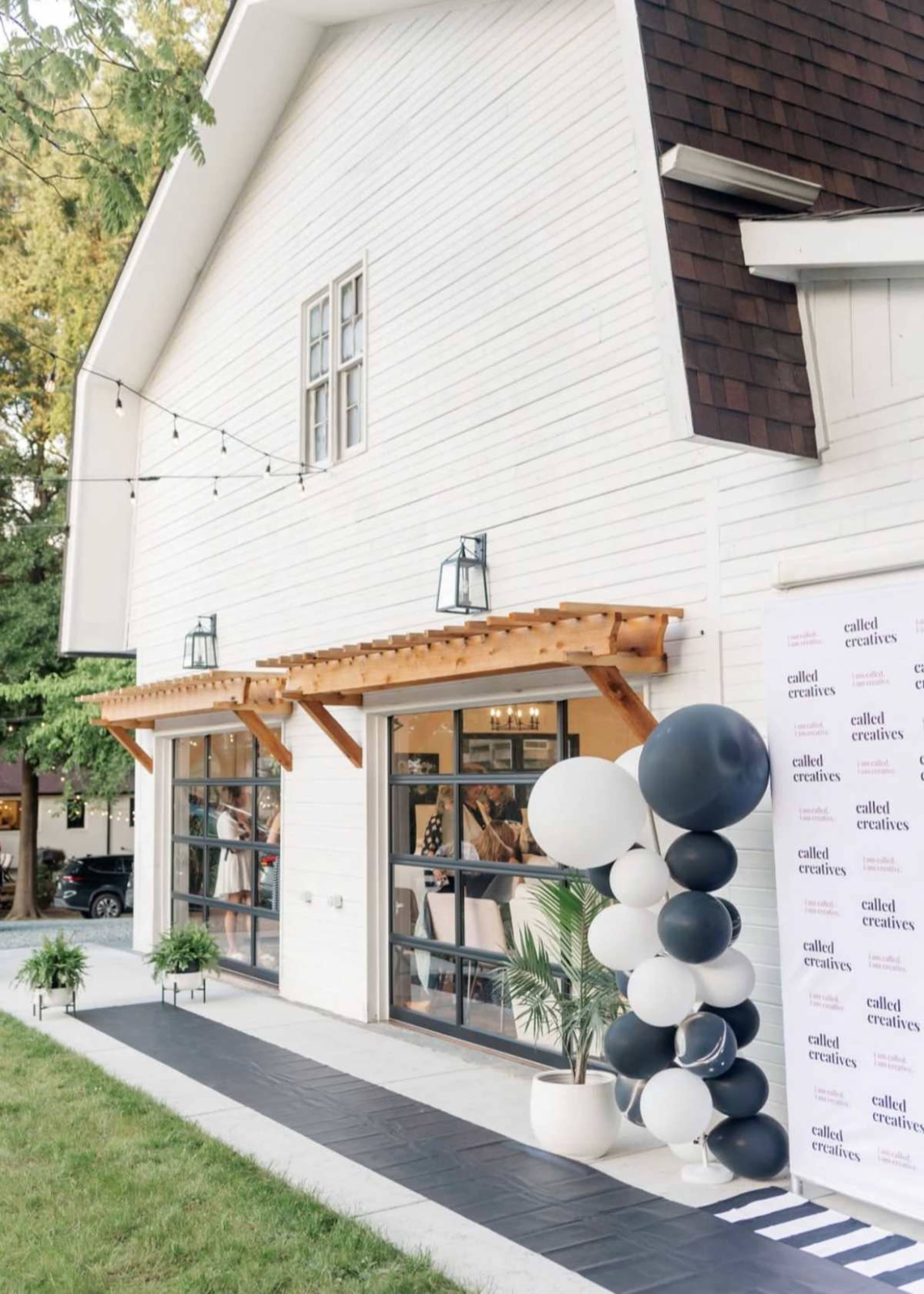 The image shows a white barn-style building with large glass doors, decorated with a balloon arch in black and white, and a pathway lined with plants leading to the entrance.