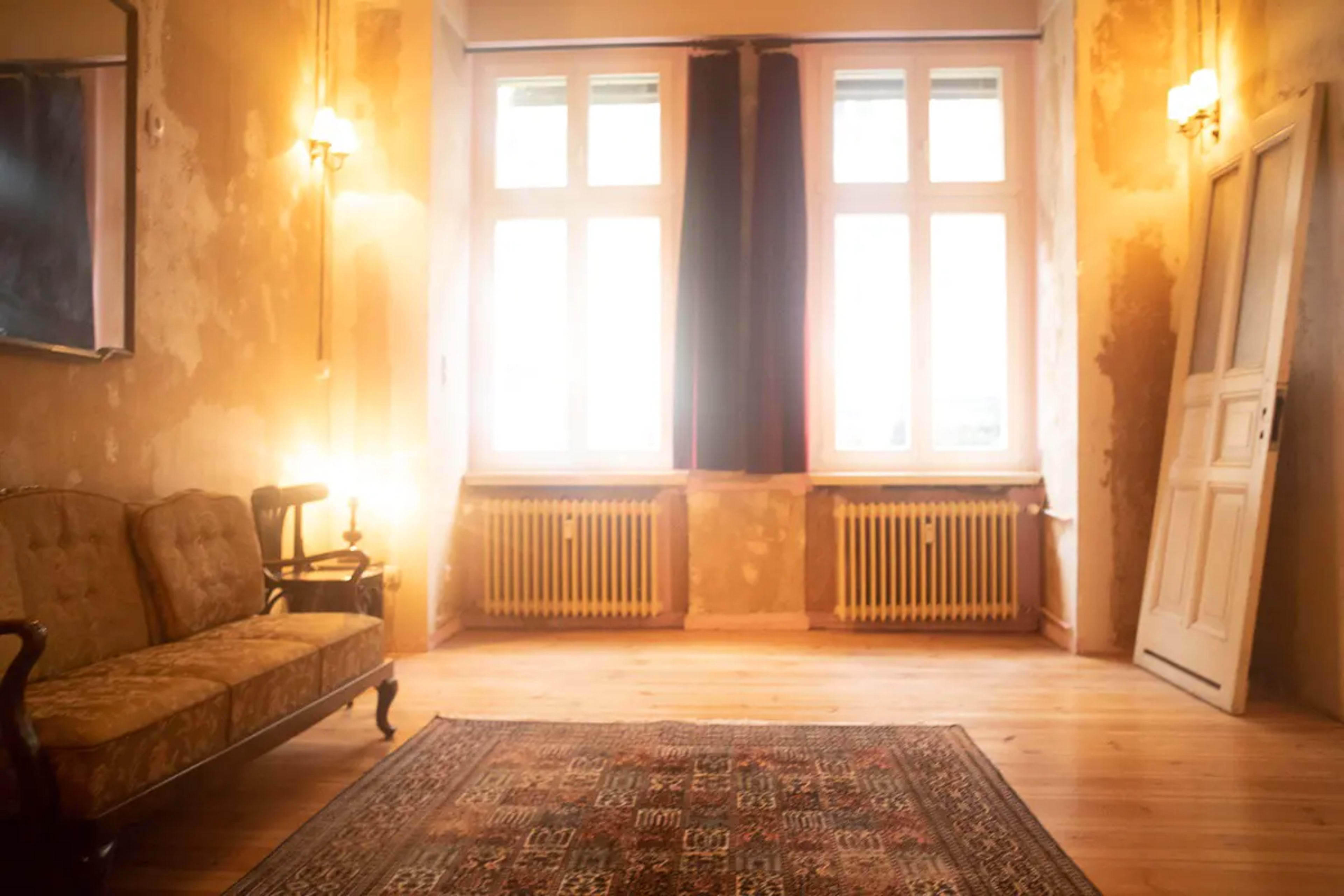A sparsely furnished room with large windows, exposed walls, a patterned rug, a vintage couch, and an open door.