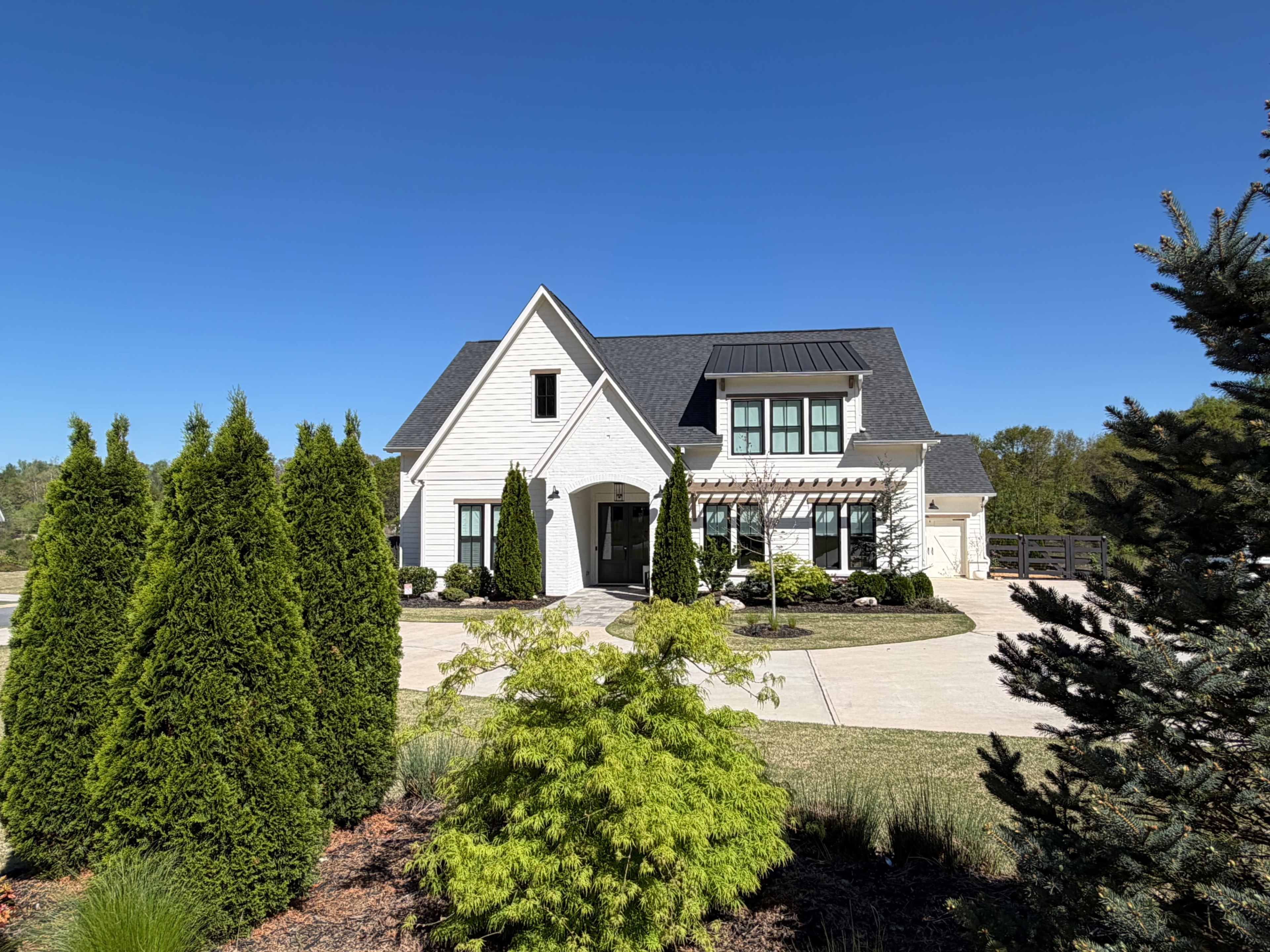 The image shows a modern two-story house with a gray roof, white siding, and neatly landscaped gardens in the foreground.