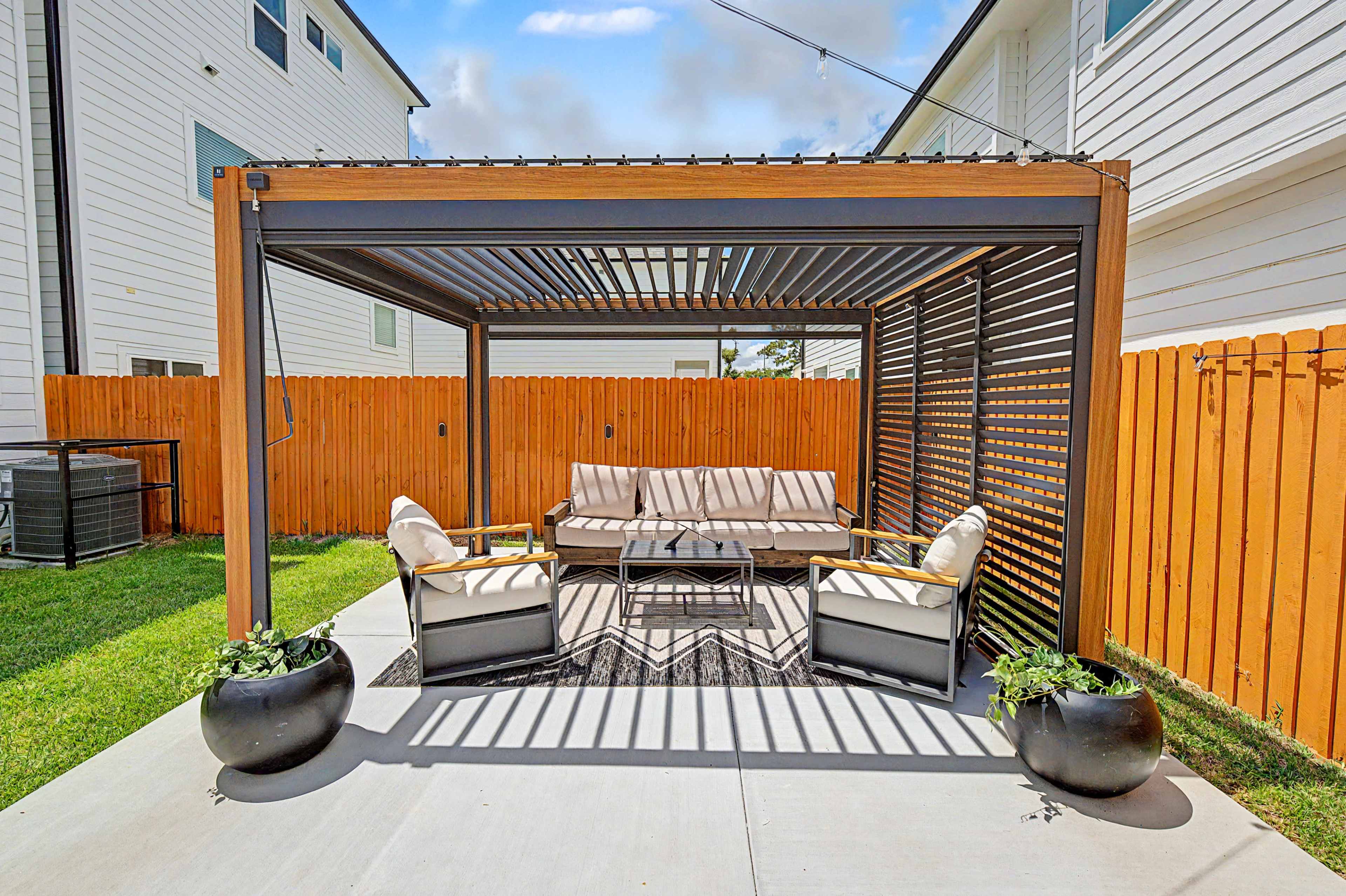 The image shows a covered outdoor seating area with a sofa and chairs, surrounded by a grassy yard and a wooden fence.