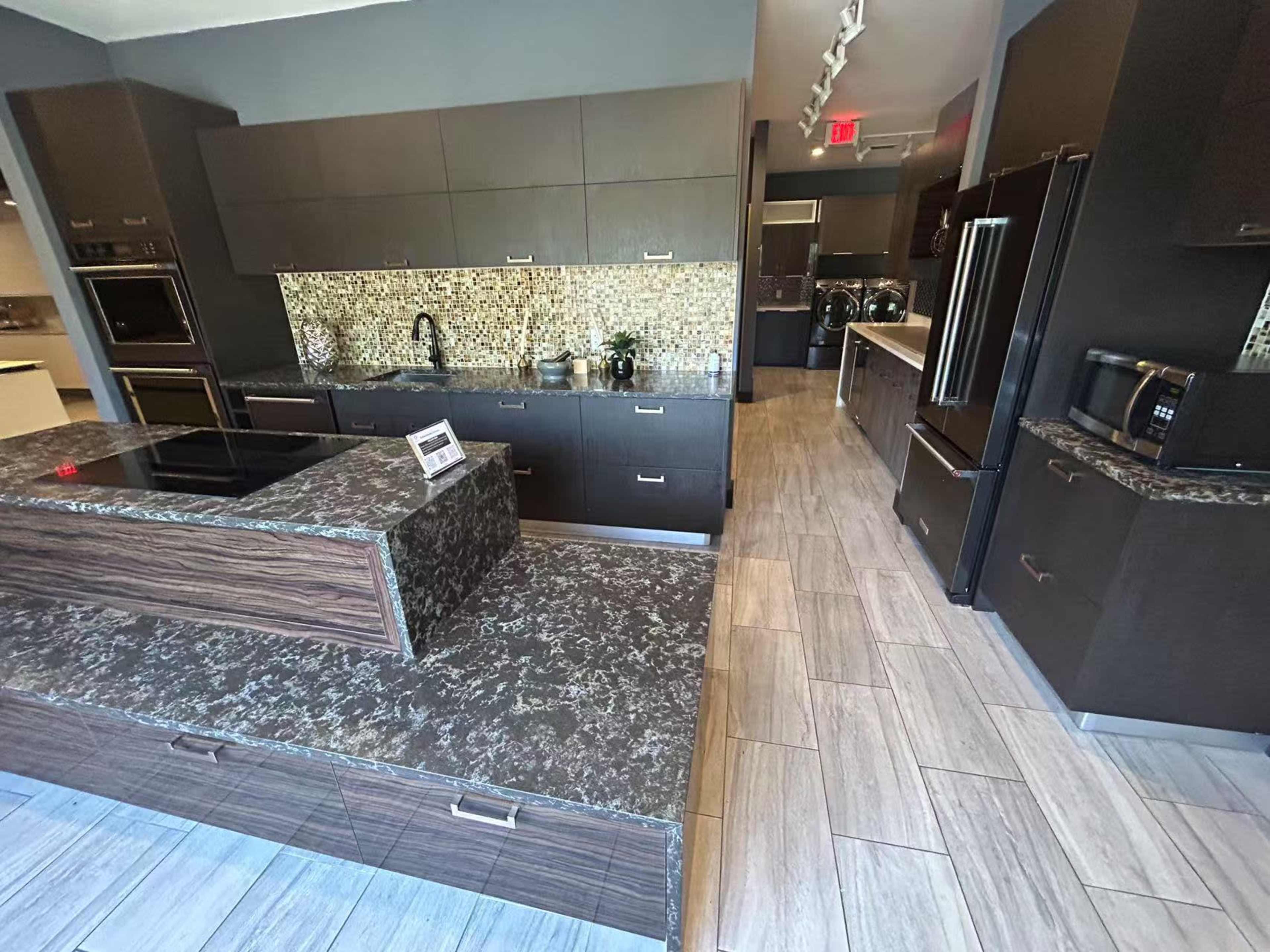 The image shows a modern kitchen featuring dark cabinetry, a central island with a stone countertop, and a tiled backsplash with a mosaic design.