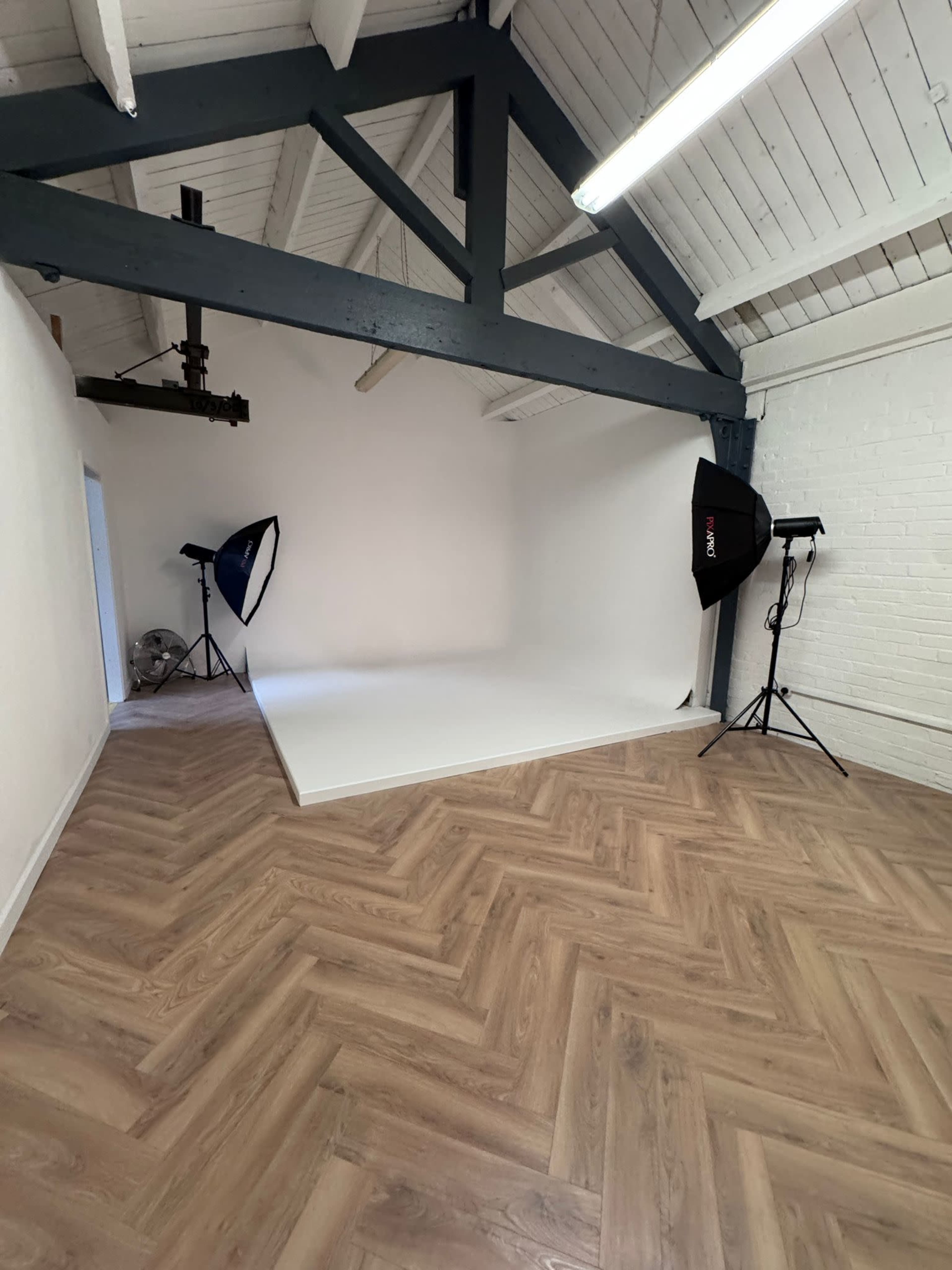 The image shows an empty studio space with a white backdrop, two softbox lighting setups, and wooden herringbone flooring.