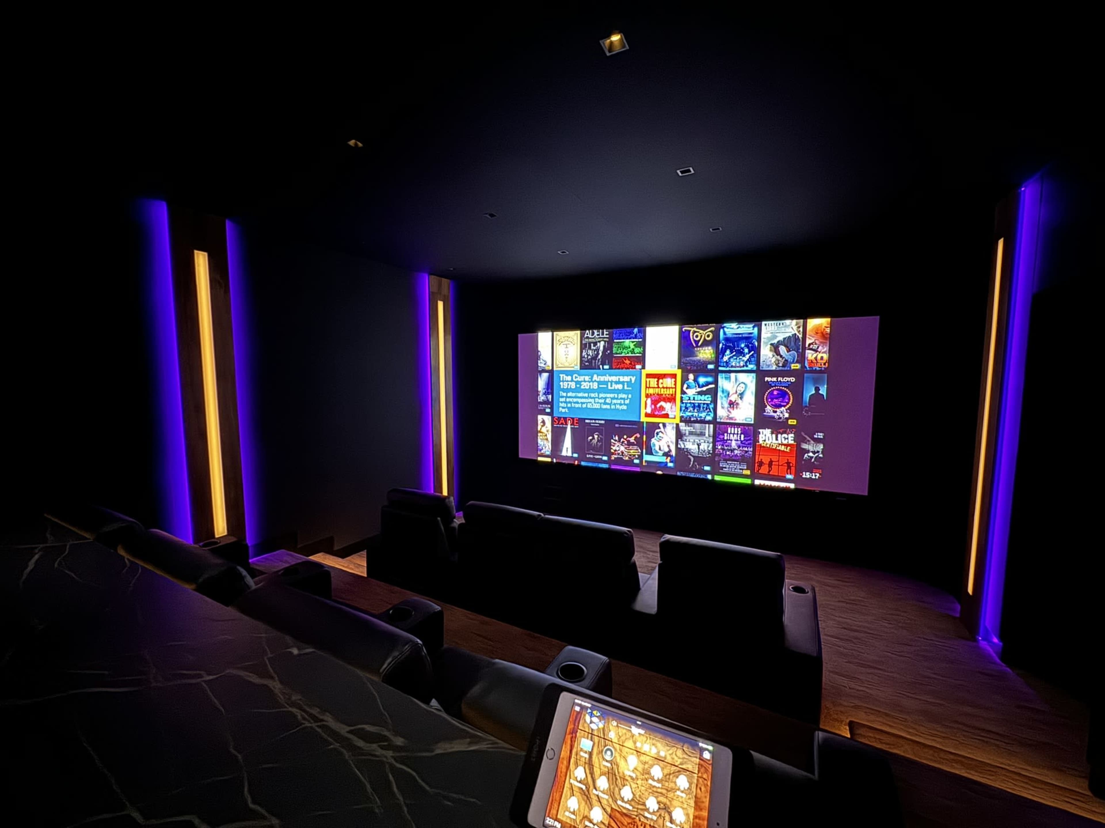 A dark home theater features a large screen displaying a movie selection interface, with leather seating and ambient lighting.