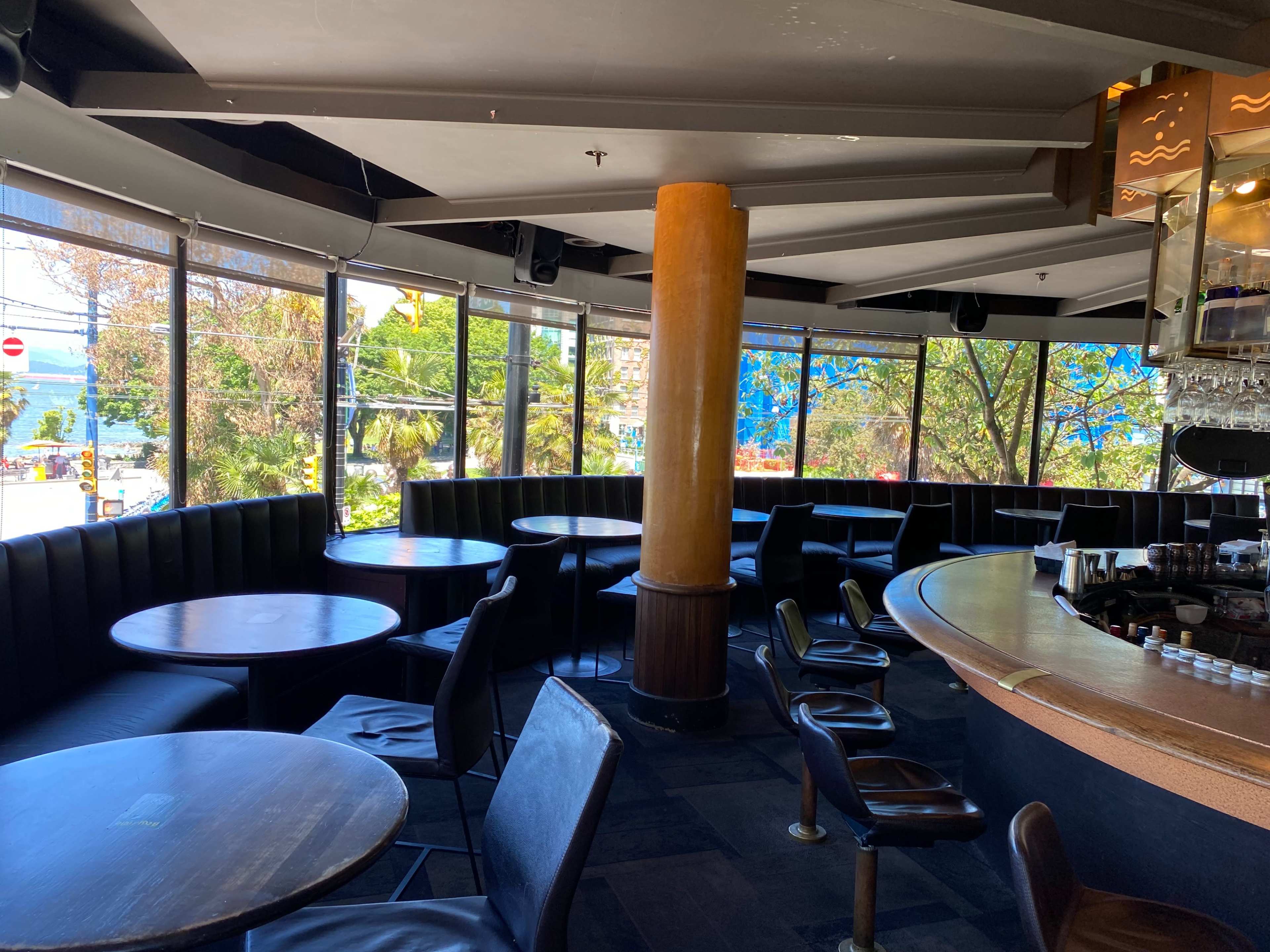 An empty bar area featuring round tables, black leather seating, and large windows overlooking a sunny outdoor scene.