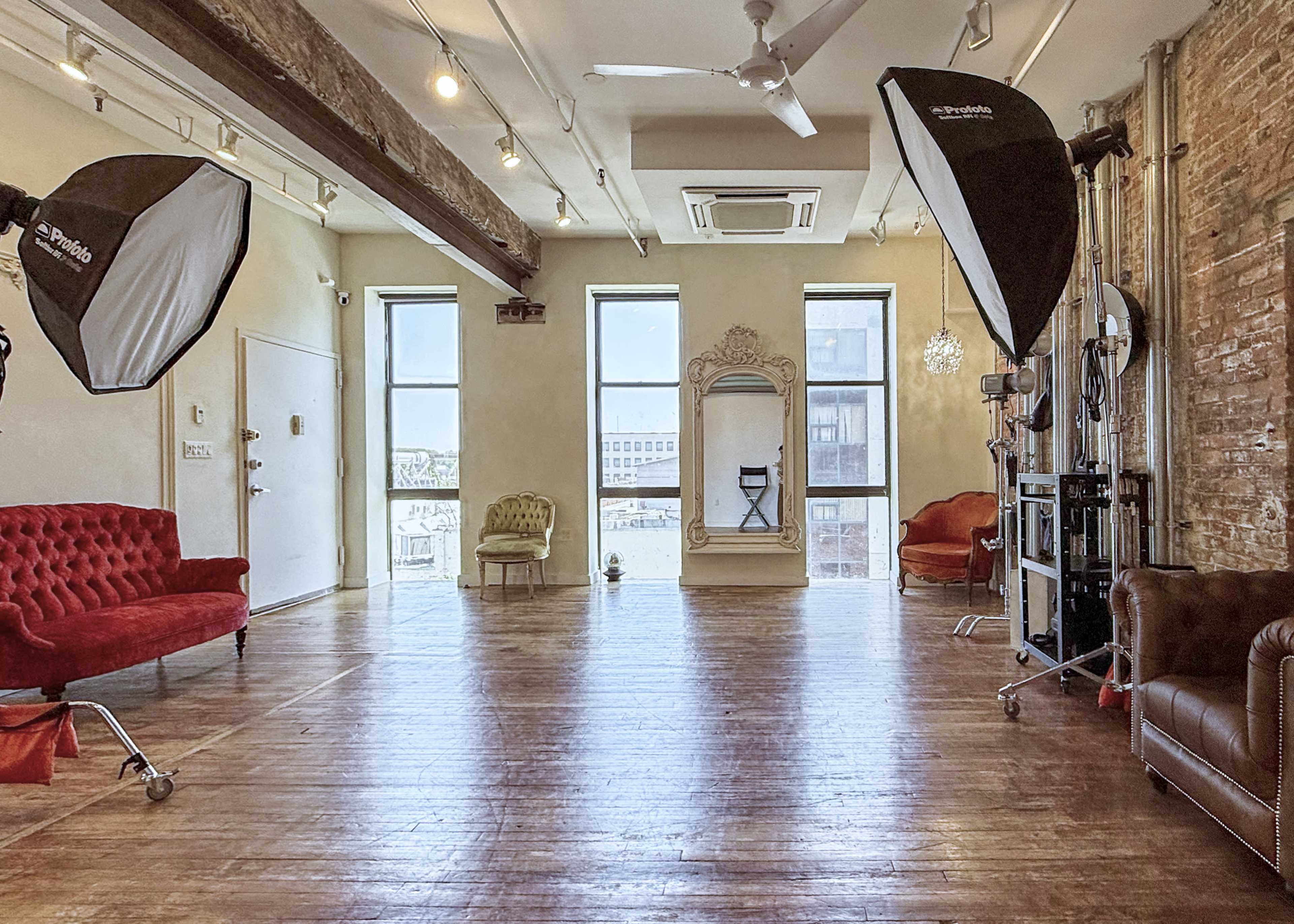 The image shows a spacious, well-lit photography studio featuring a mix of seating options, large windows, and professional lighting equipment.