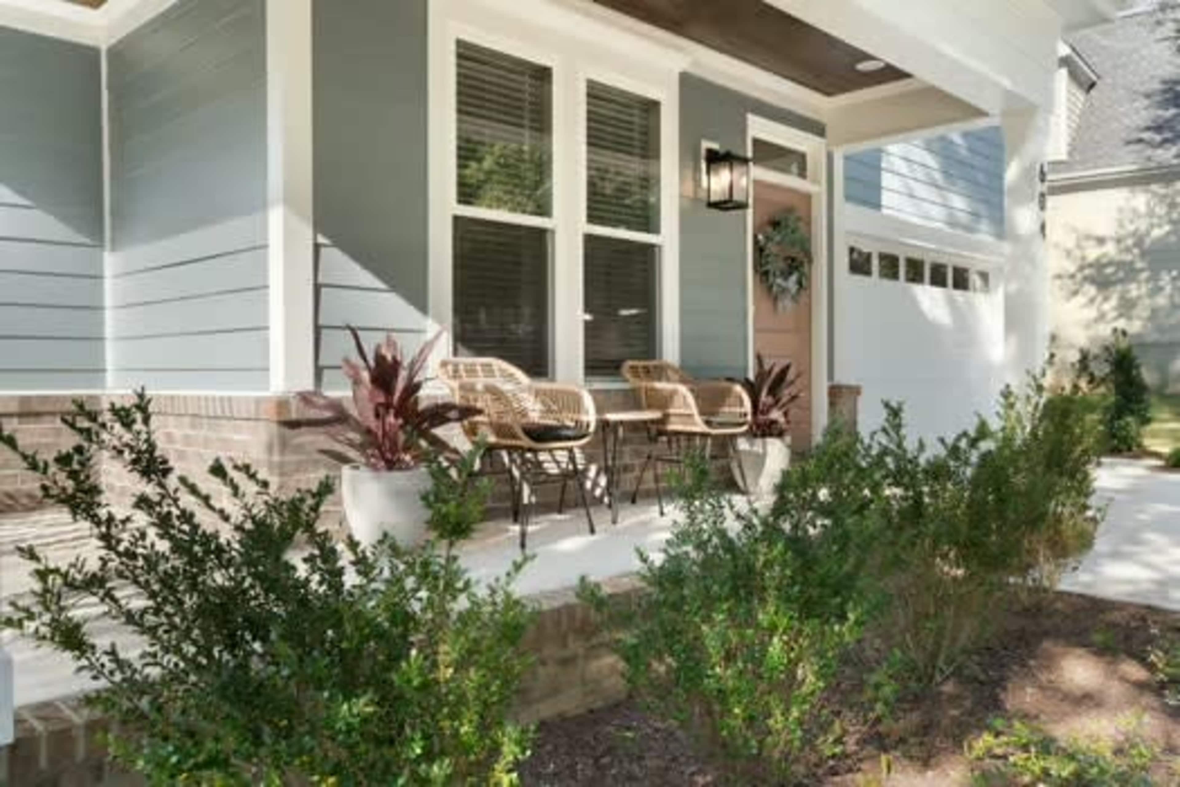 The image shows a front porch with two chairs, a small side table, and potted plants, surrounded by a well-maintained landscape.