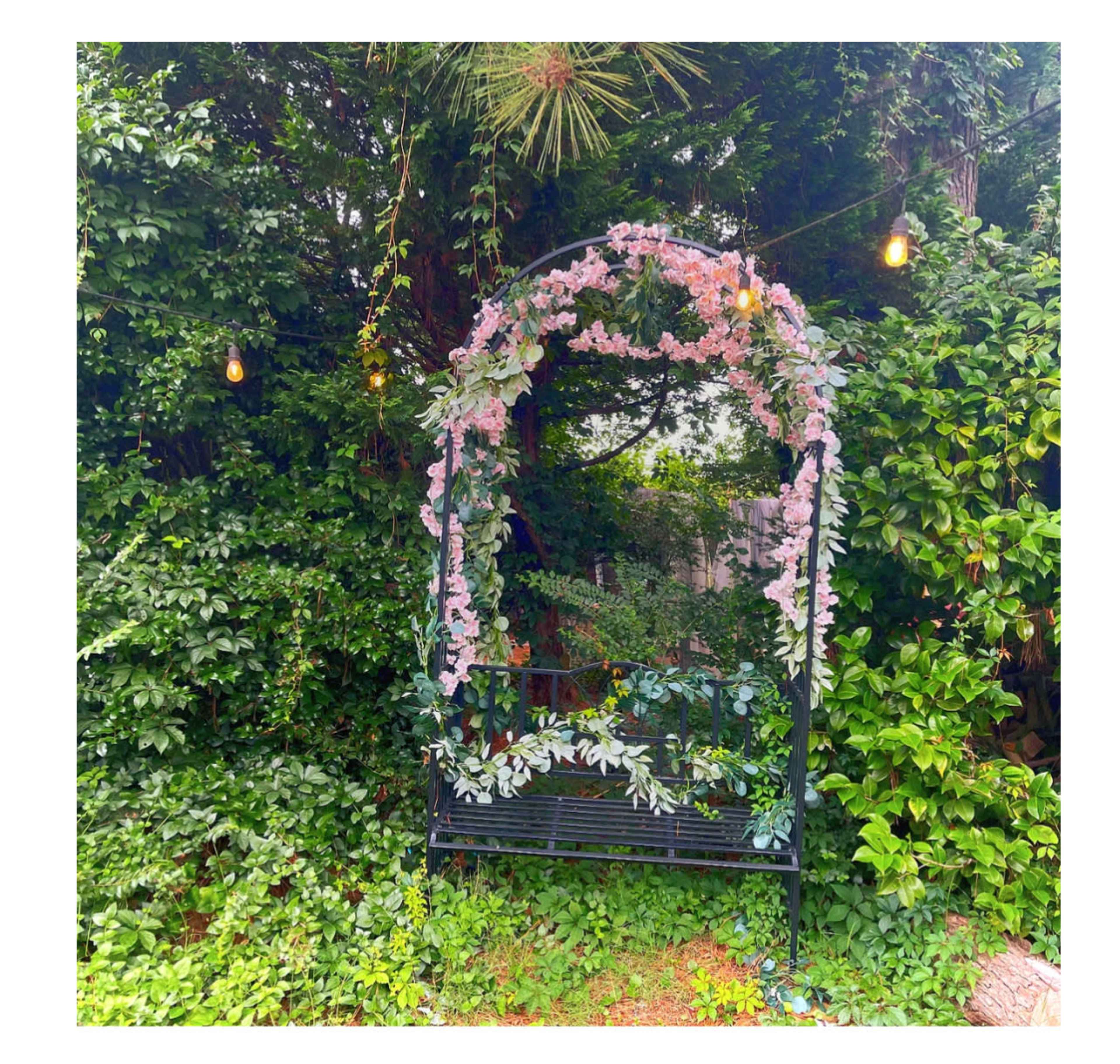 A metal bench is partially covered by greenery and adorned with pink flowers, set against a dense backdrop of foliage and hanging lights.