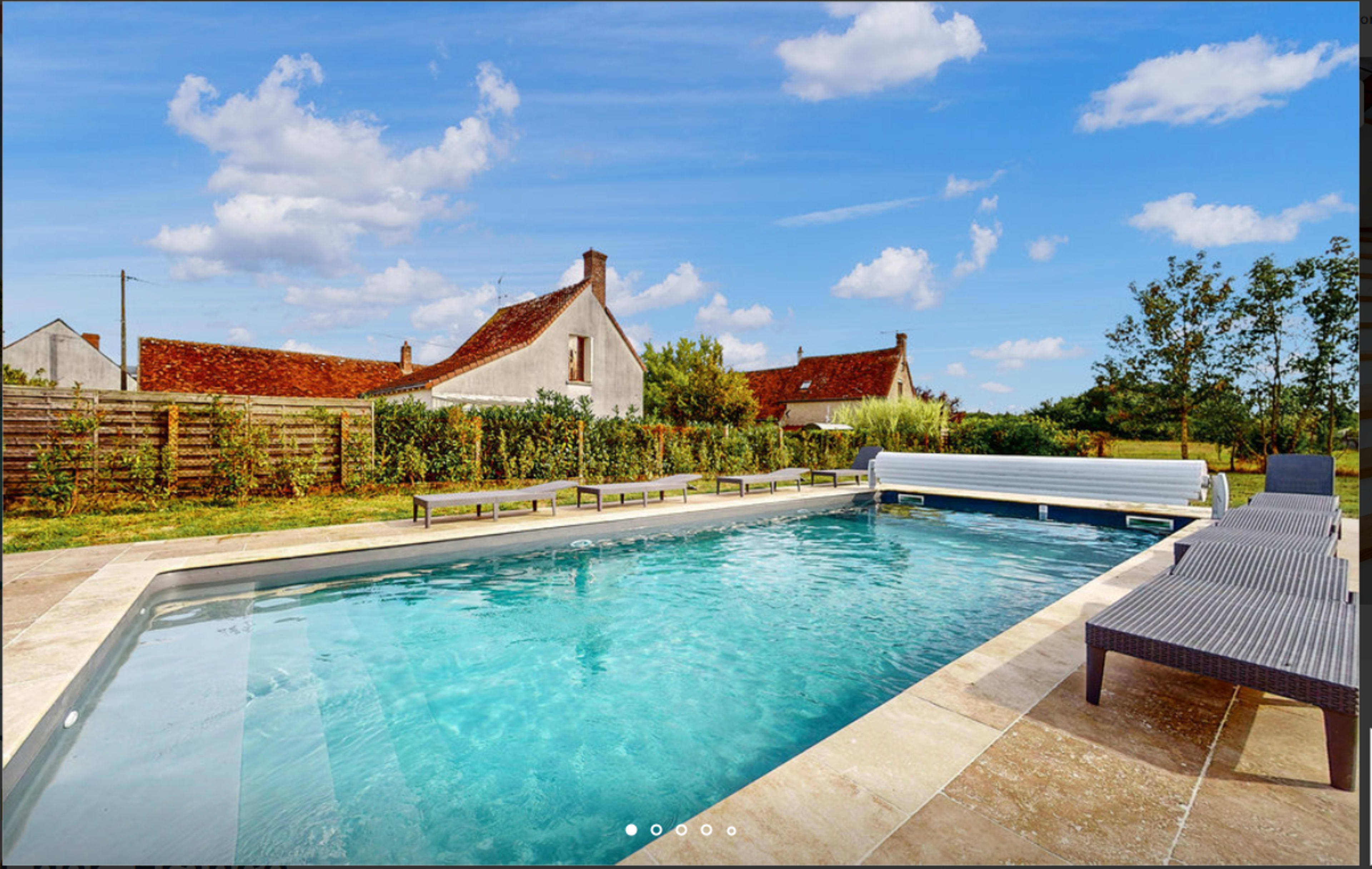 The image shows a clear swimming pool surrounded by a patio with loungers and a backdrop of rustic houses and greenery under a blue sky with scattered clouds.
