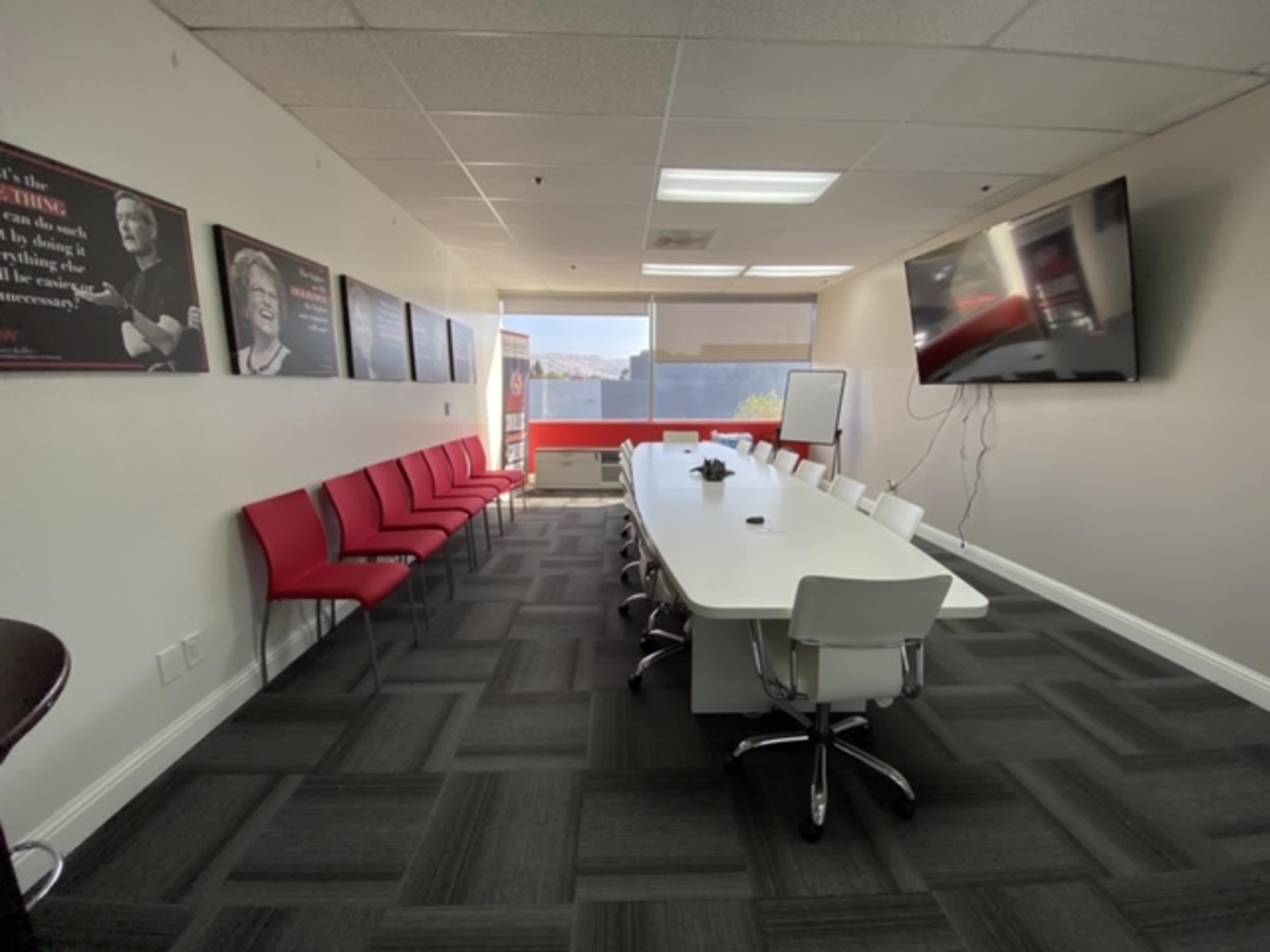 The image shows a modern conference room with a long white table, several chairs, and a large wall-mounted television.