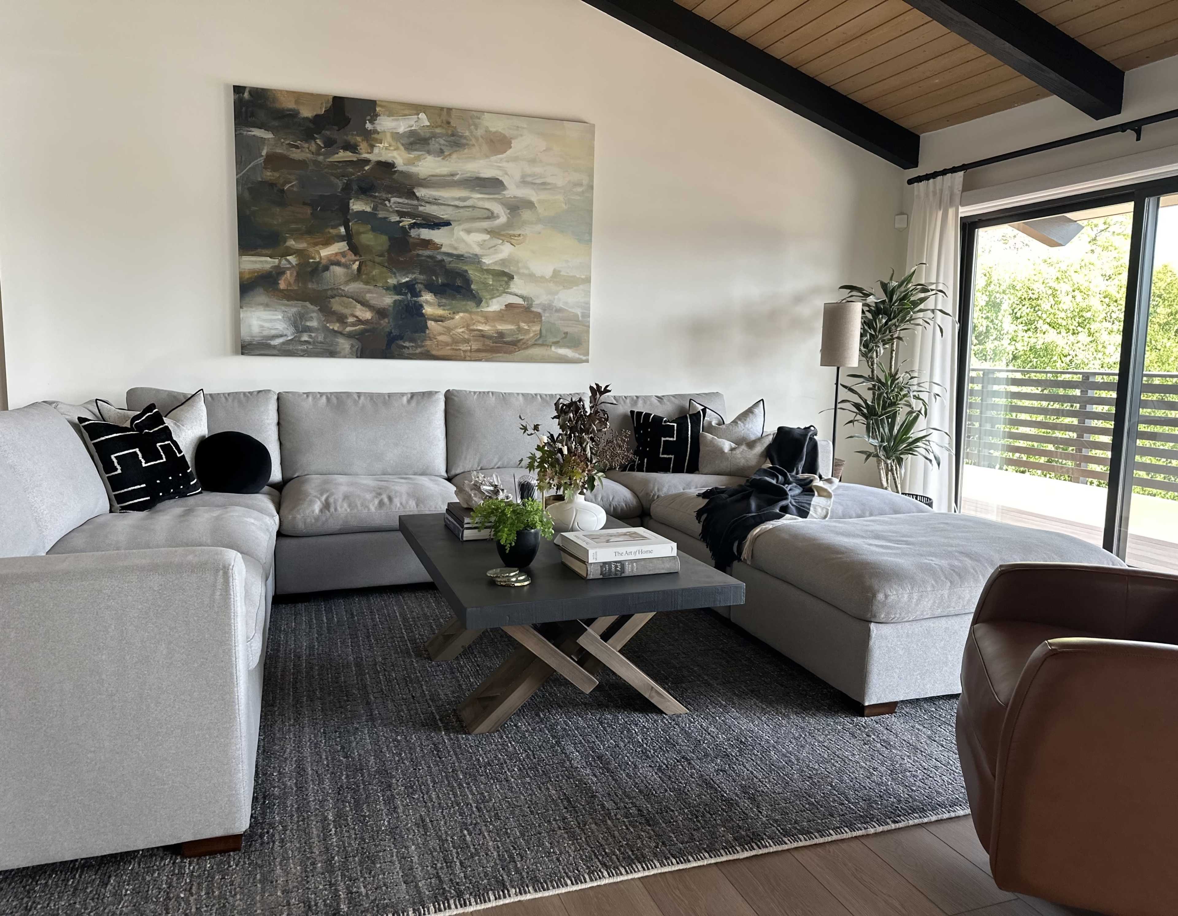 A modern living room features a light gray sectional sofa, a central coffee table with a decorative arrangement, and large windows opening to a balcony with greenery outside.
