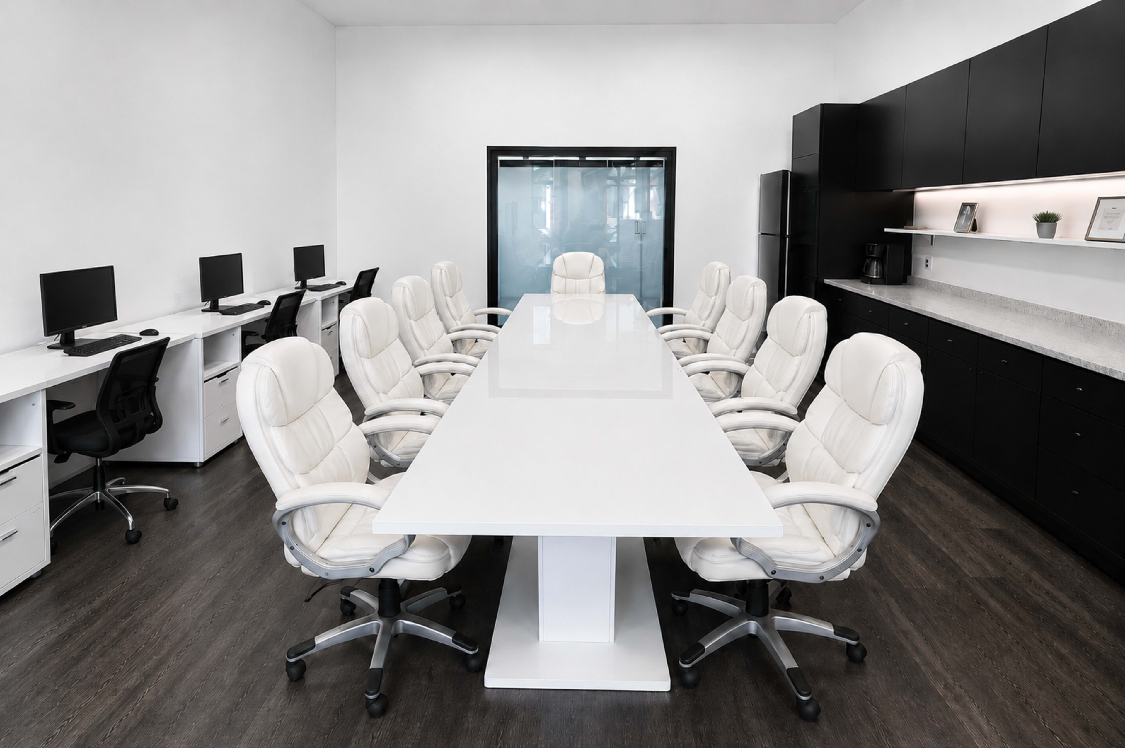 The image depicts a modern conference room with a large white table surrounded by sixteen white office chairs, flanked by workstations with computers along one wall and a black kitchenette along the opposite wall.