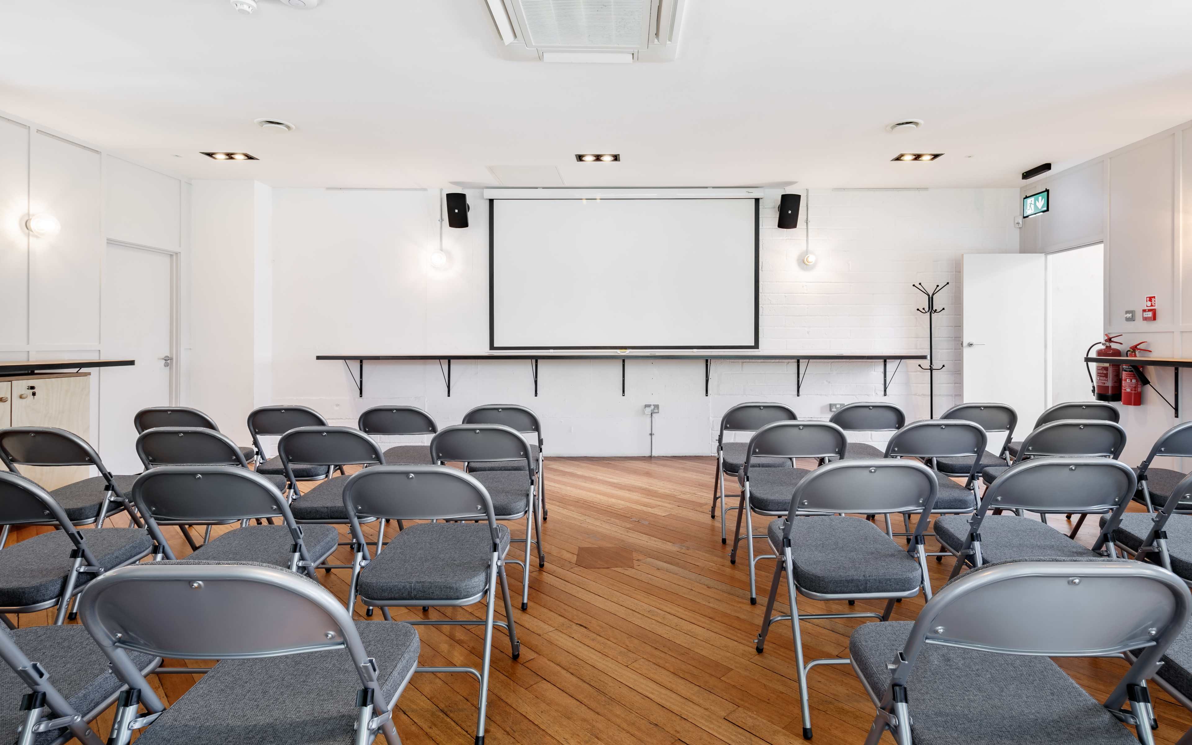 A conference room is set up with rows of gray chairs facing a wall-mounted projector screen.