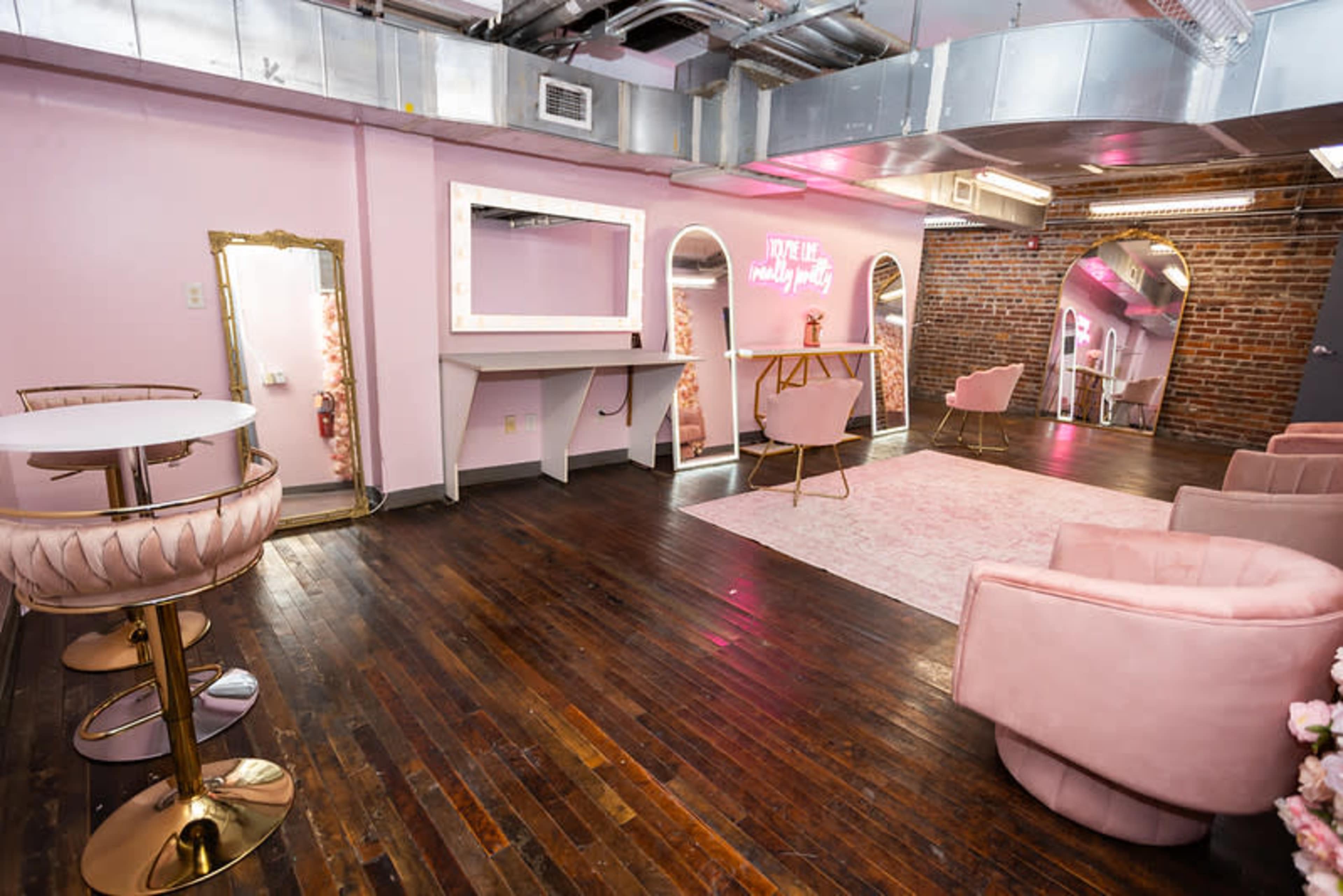 The image shows a stylish, pink-themed lounge area with a wooden floor, featuring seating, a mirror, and decorative neon signage.
