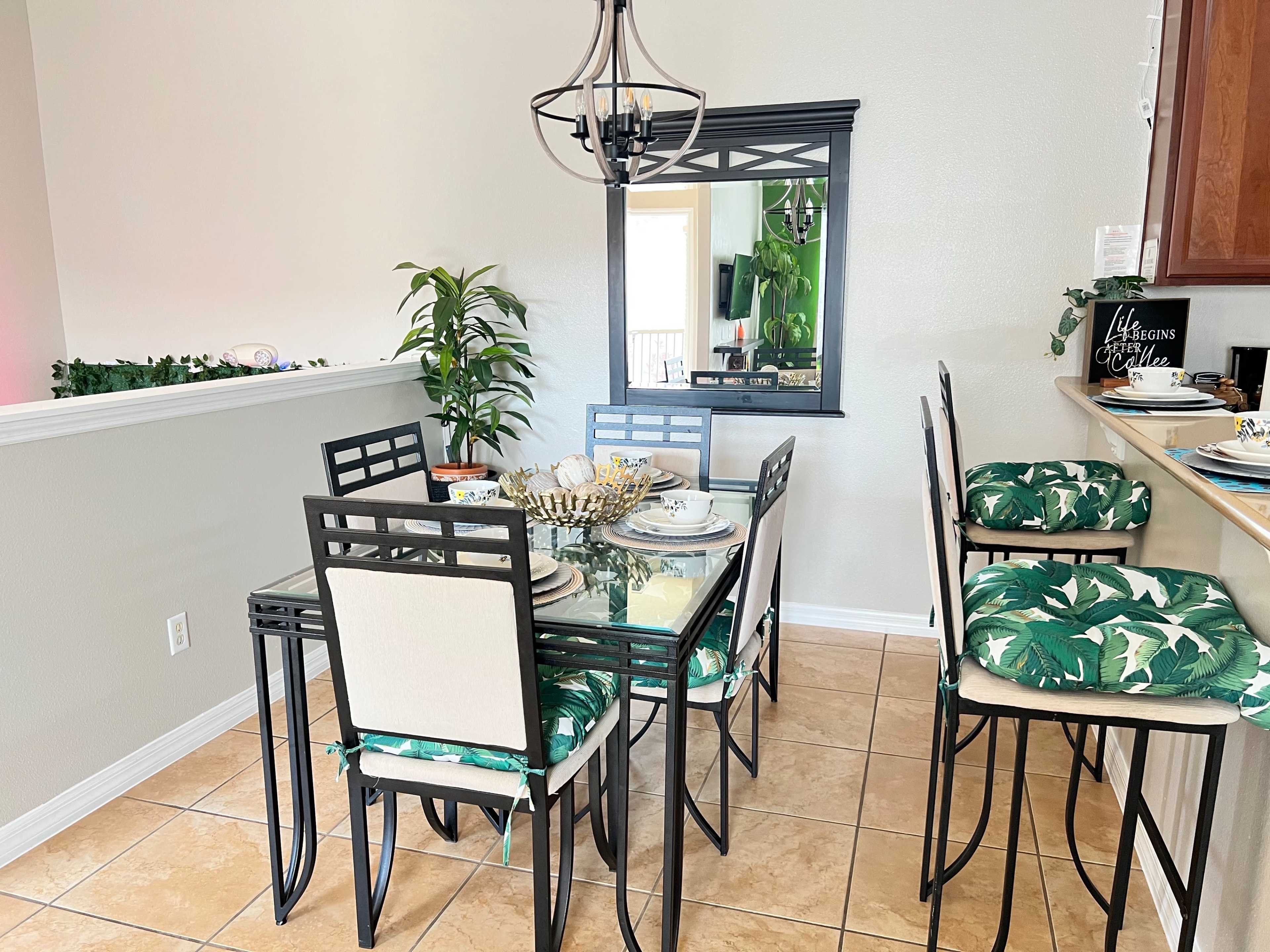 The image shows a dining area with a glass-top table surrounded by six chairs, adorned with green tropical-patterned cushions, next to a wall-mounted mirror and a potted plant.