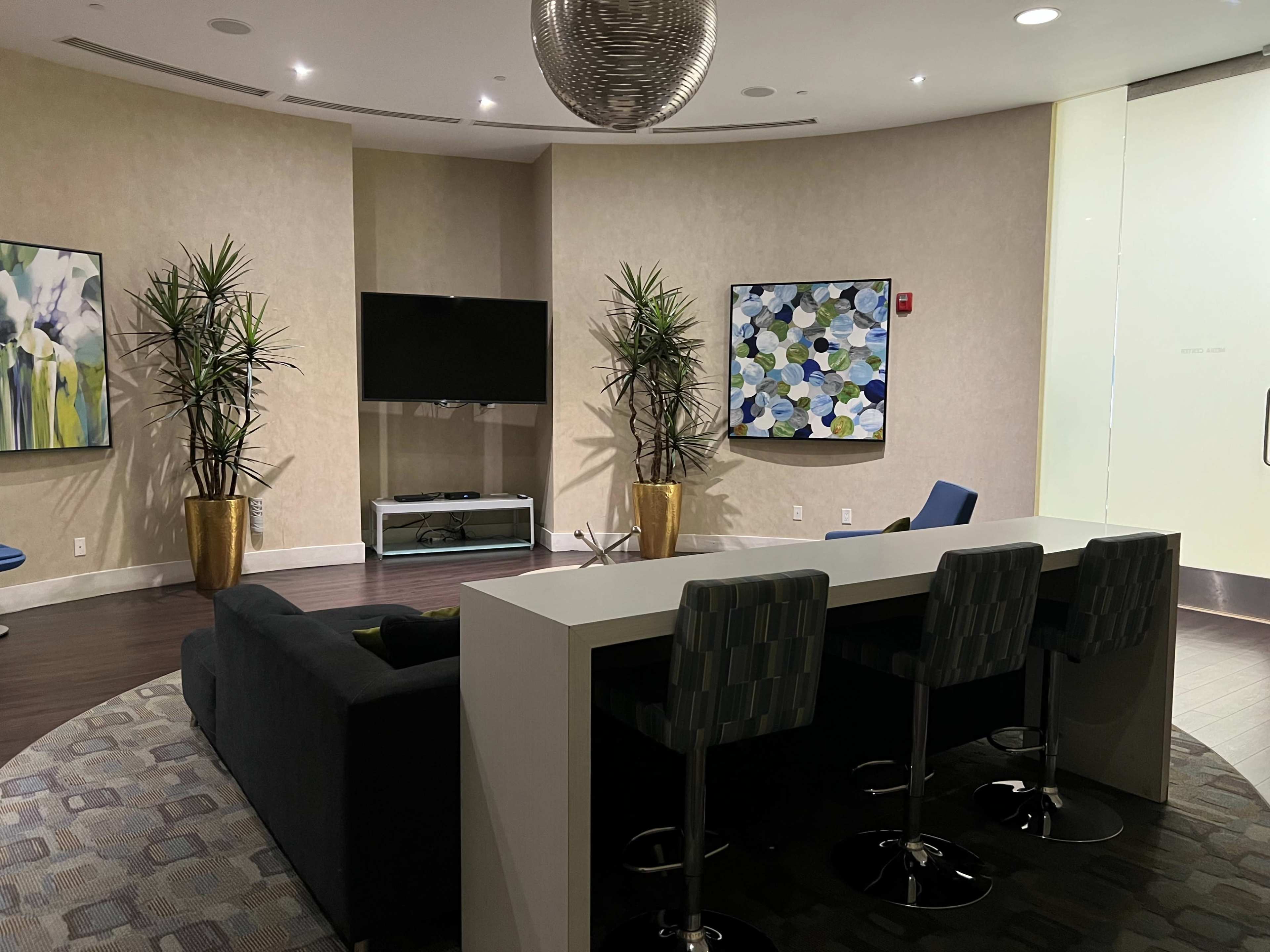 A modern lounge area with a dark sofa, a flat-screen TV, two decorative paintings, and a kitchen counter with four stools.