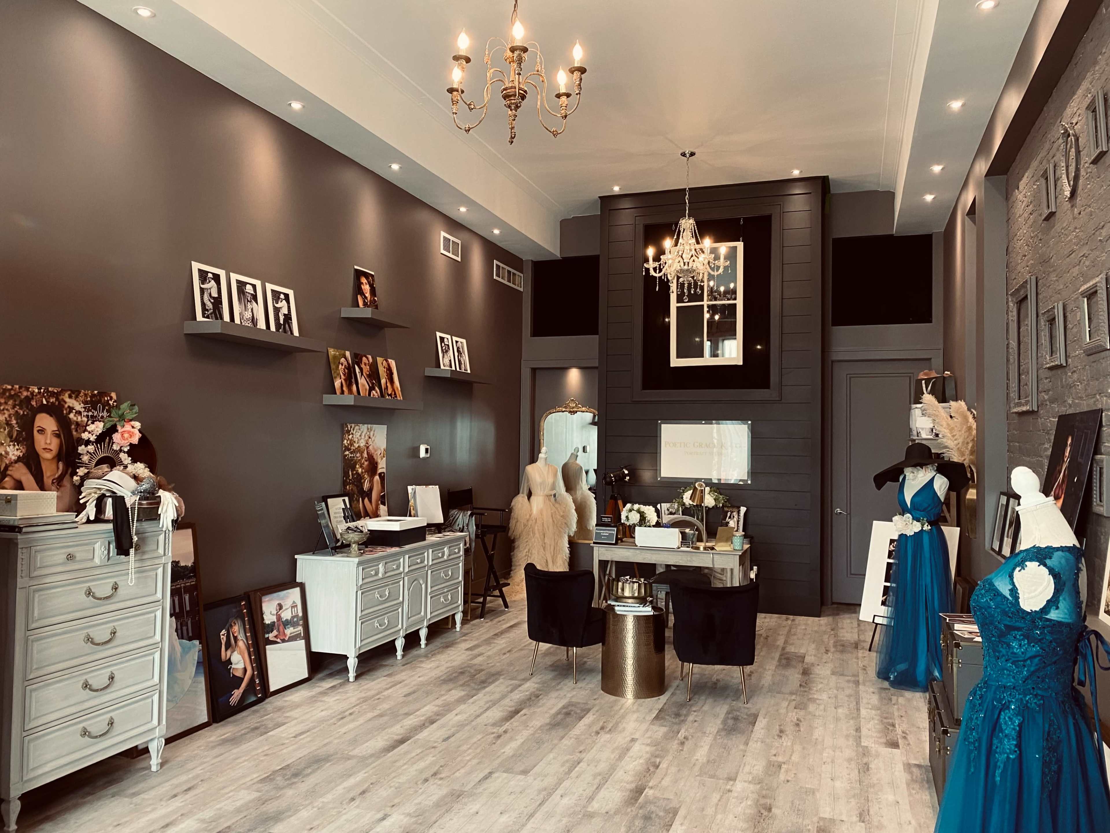 The interior of a boutique features elegant displays of dresses and framed photographs against a dark wall, illuminated by a chandelier.