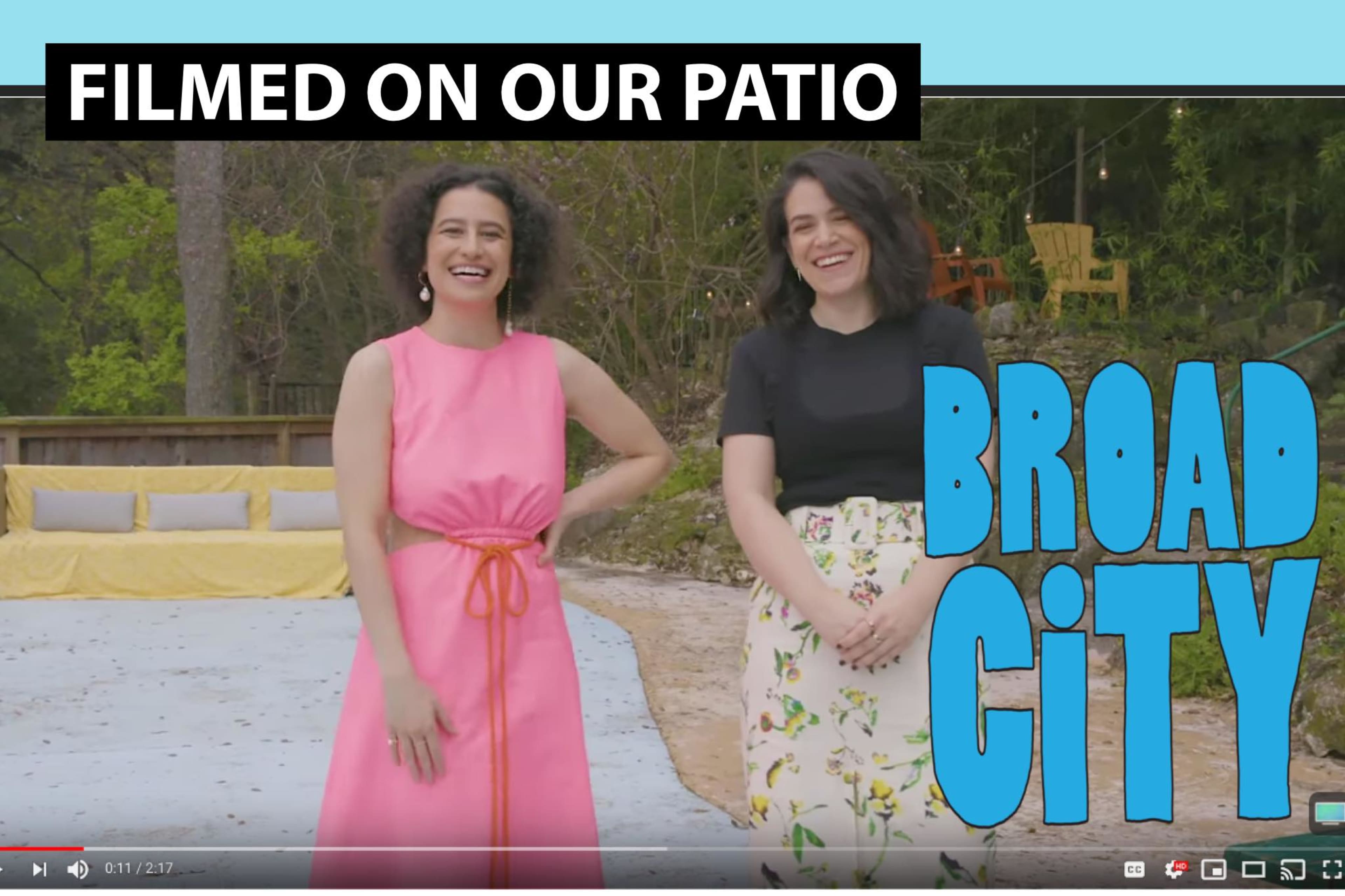 Two women stand smiling on a patio, one in a pink dress and the other in a black shirt and floral skirt, with "Broad City" displayed in bold blue lettering.