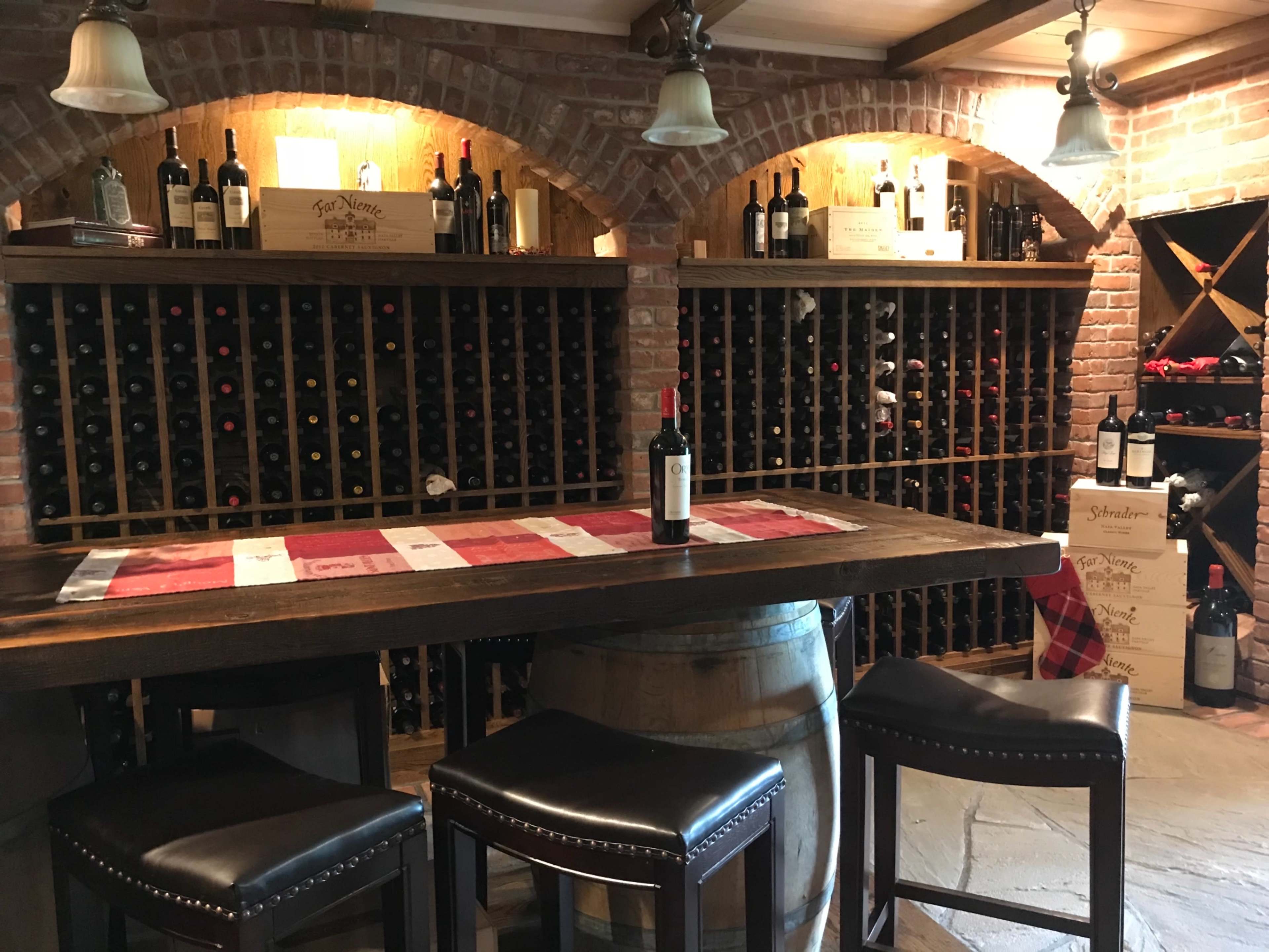A rustic wine cellar featuring wooden wine racks filled with bottles, a bar table set with a wine bottle, and high-backed stools.