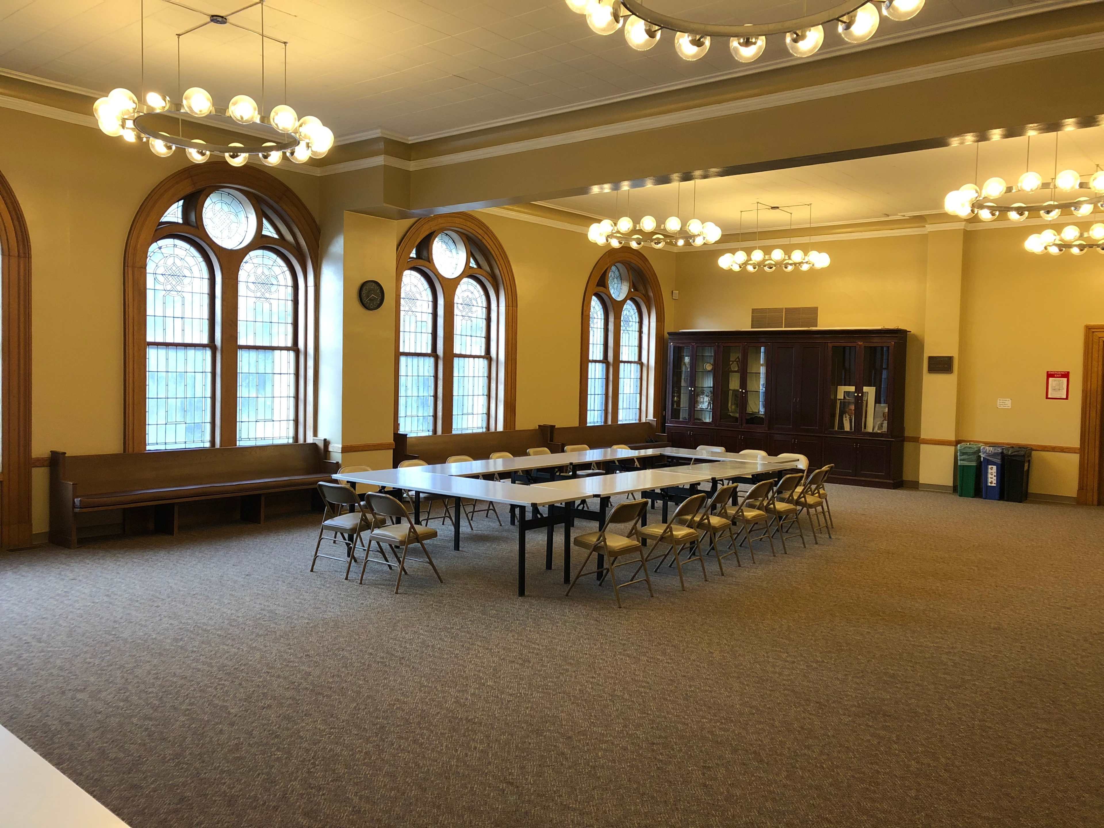 A spacious room features arched windows, a long table surrounded by chairs, and a dark wood cabinet along one wall.