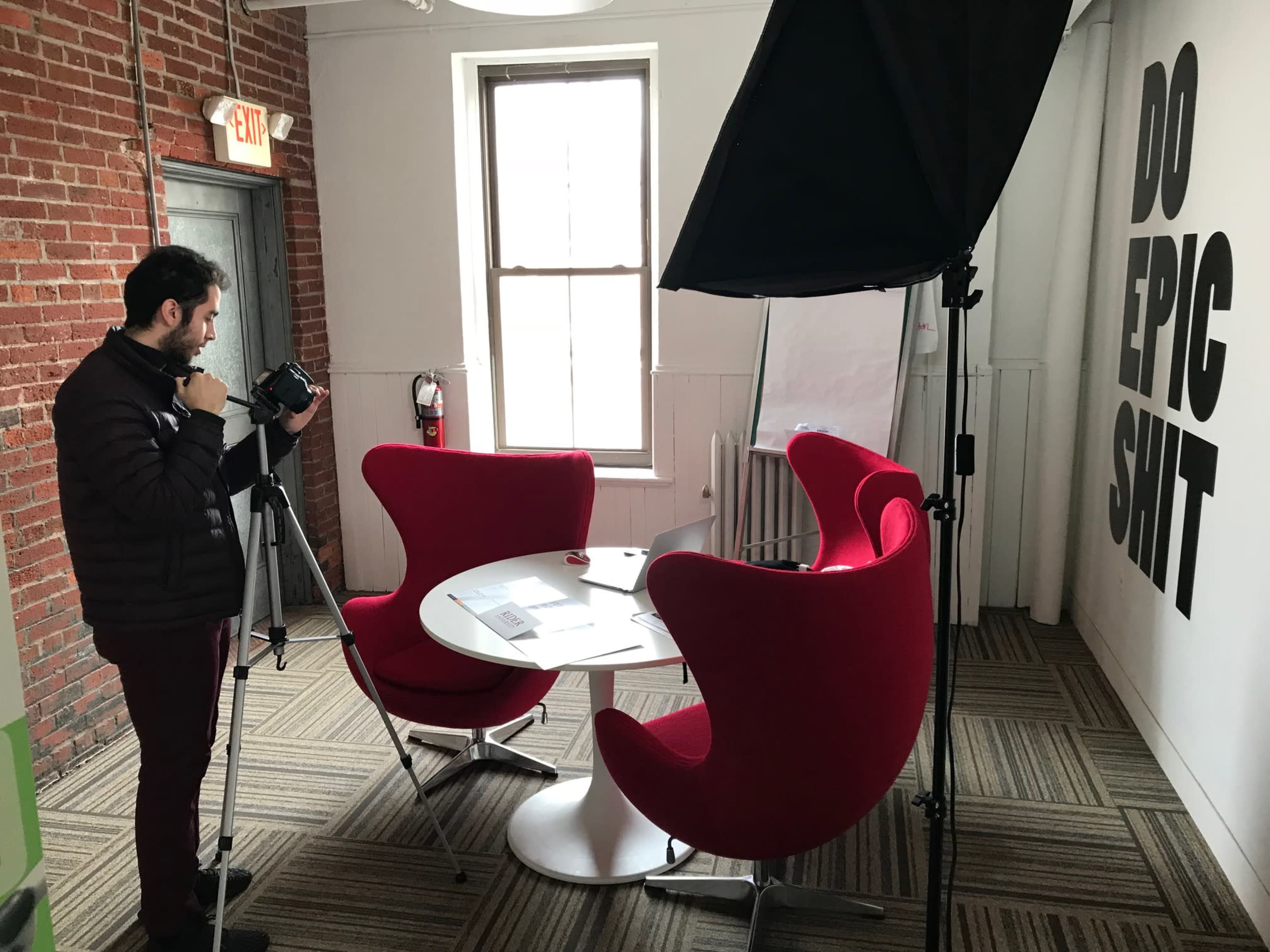 A person is setting up a camera on a tripod in a room with red chairs and a motivational message on the wall.