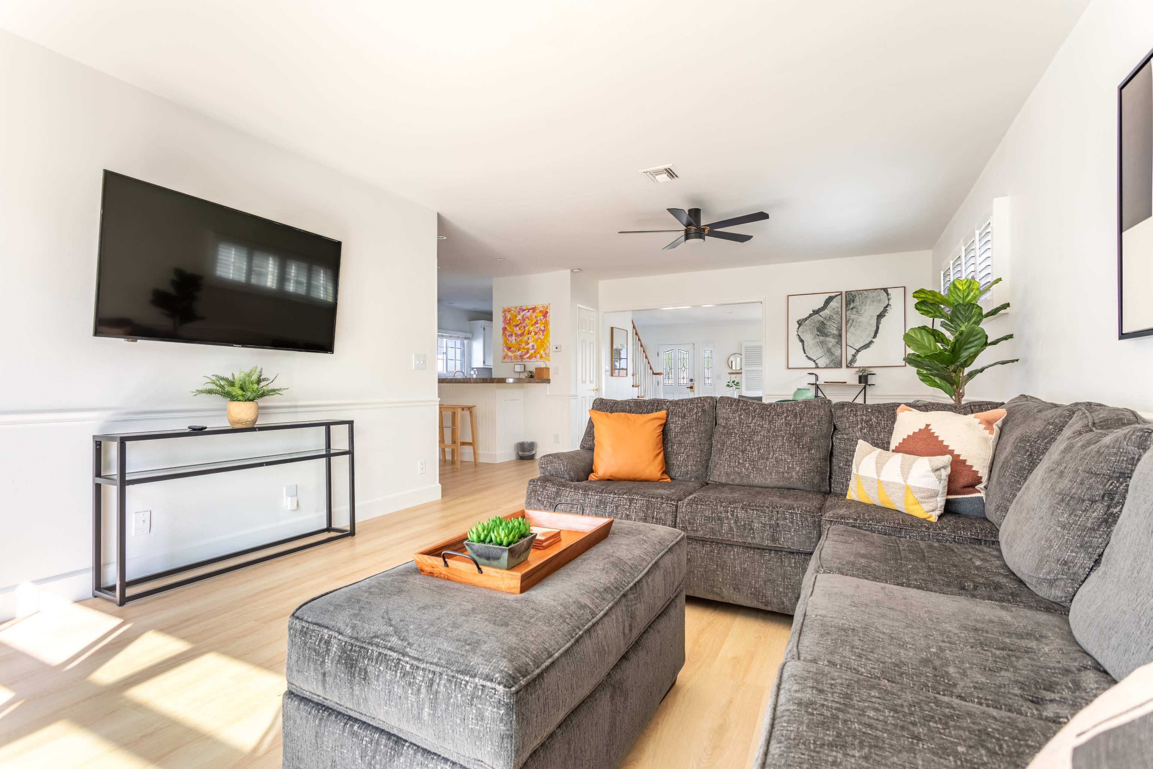 A modern living room features a gray sectional sofa, a mounted television, and a coffee table with a decorative tray, set against a bright, open space with kitchen access.