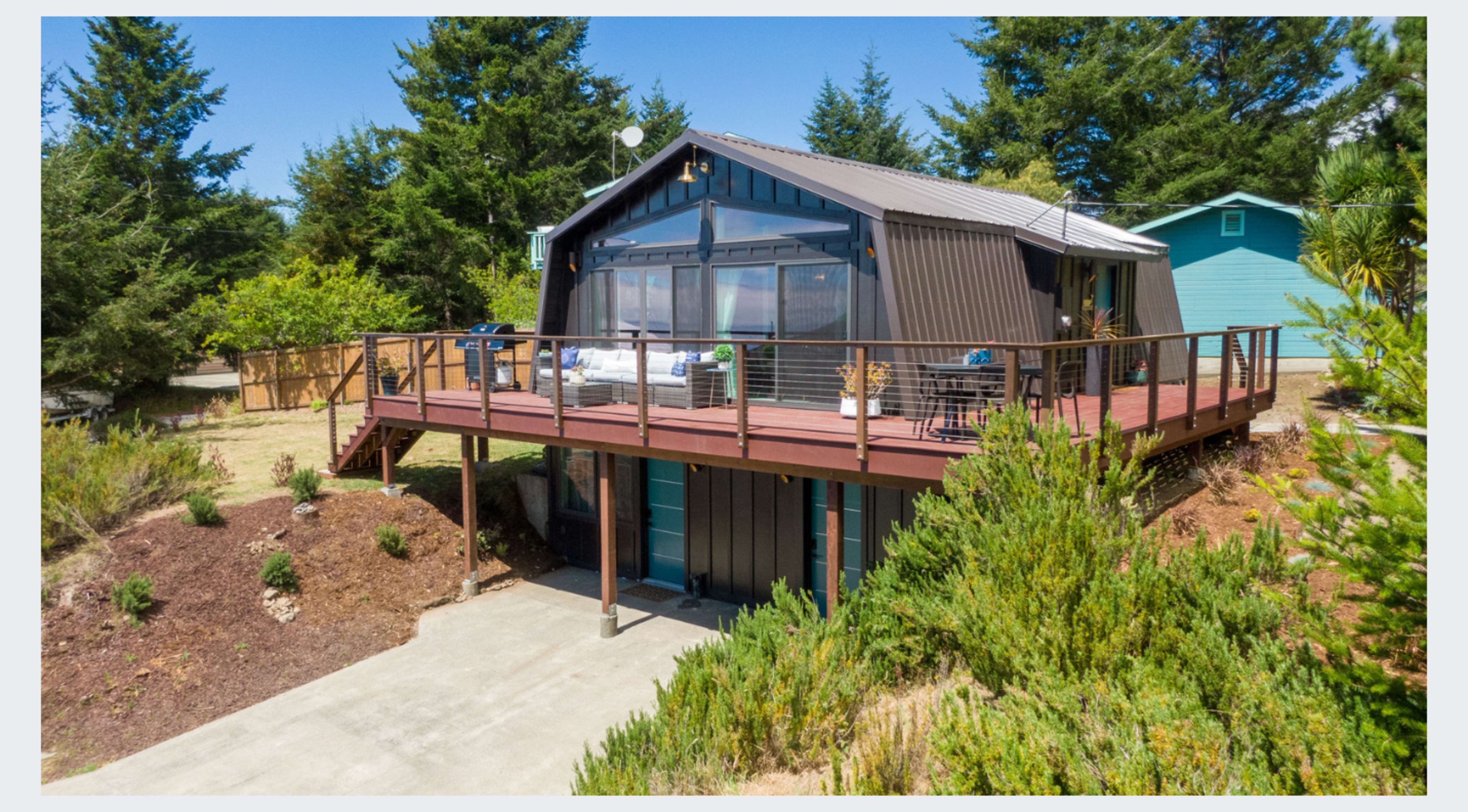 A modern house with a wooden deck is situated on a sloped landscape surrounded by greenery and tall trees.