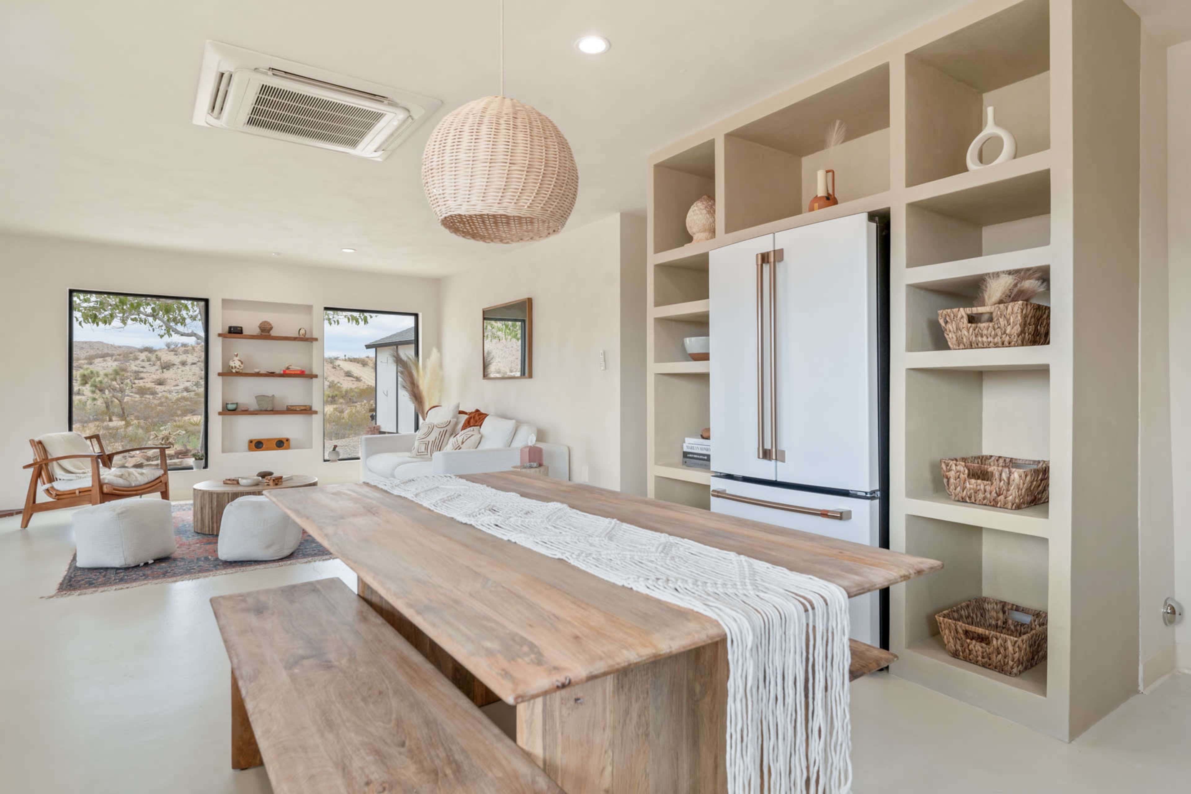 A modern open-concept living space features a wooden dining table, a white refrigerator, and shelves with decorative items, all set against a backdrop of large windows overlooking a desert landscape.