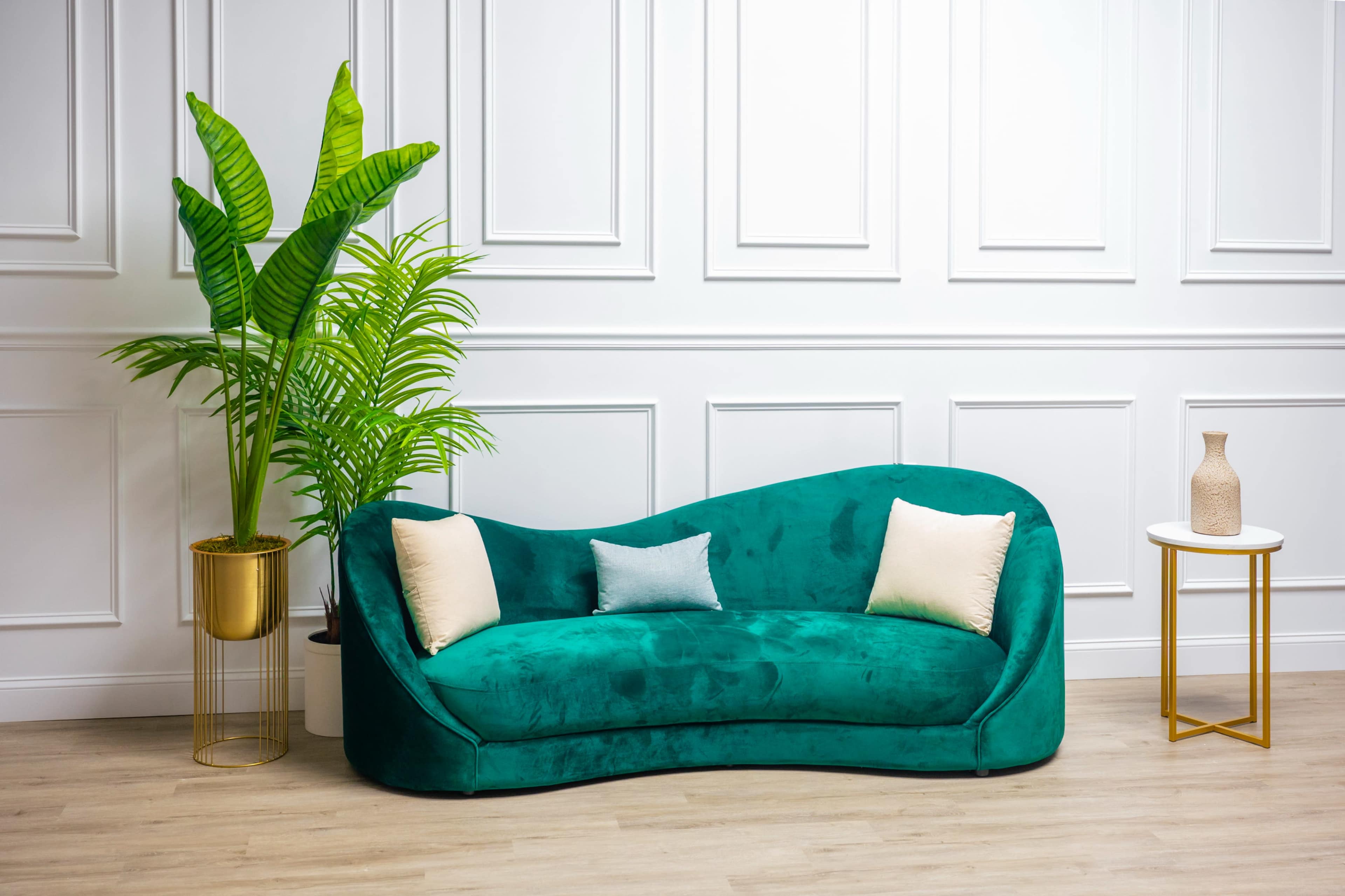 A curved, green velvet sofa with decorative cushions, accompanied by a large potted plant and a small side table.