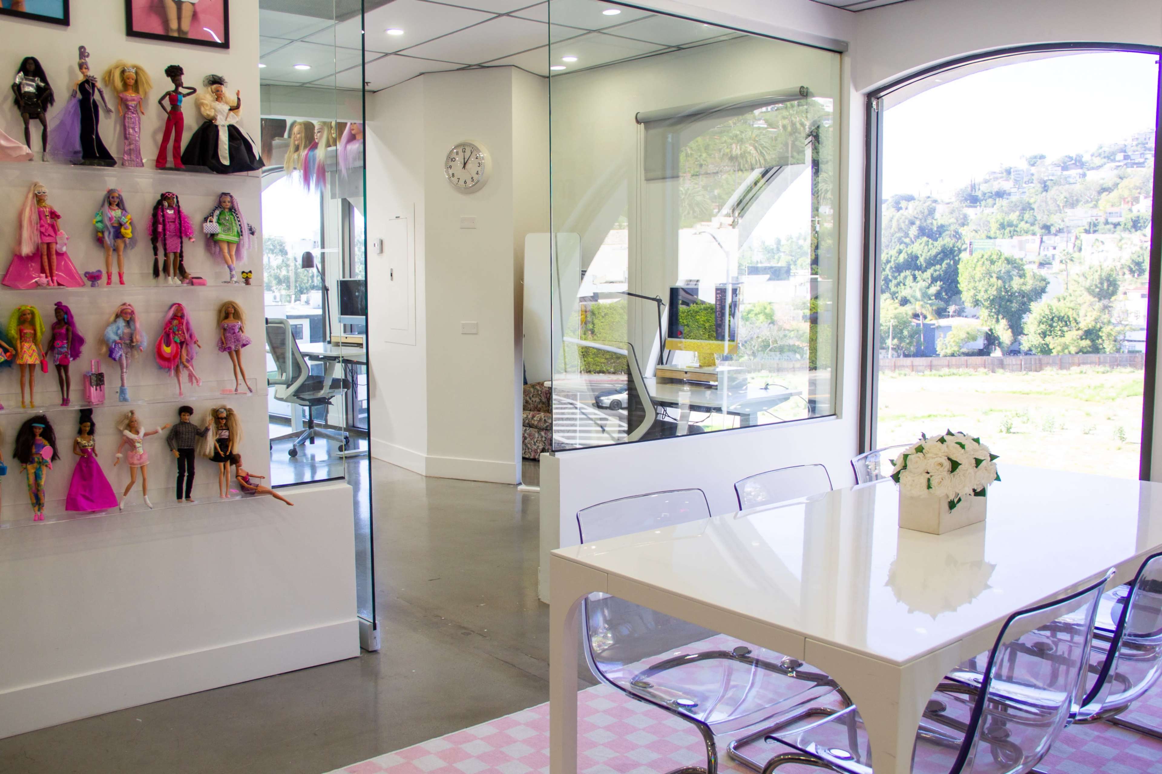 The image shows a modern office space with a clear glass wall, a white table surrounded by transparent chairs, and a display of dolls on the wall.