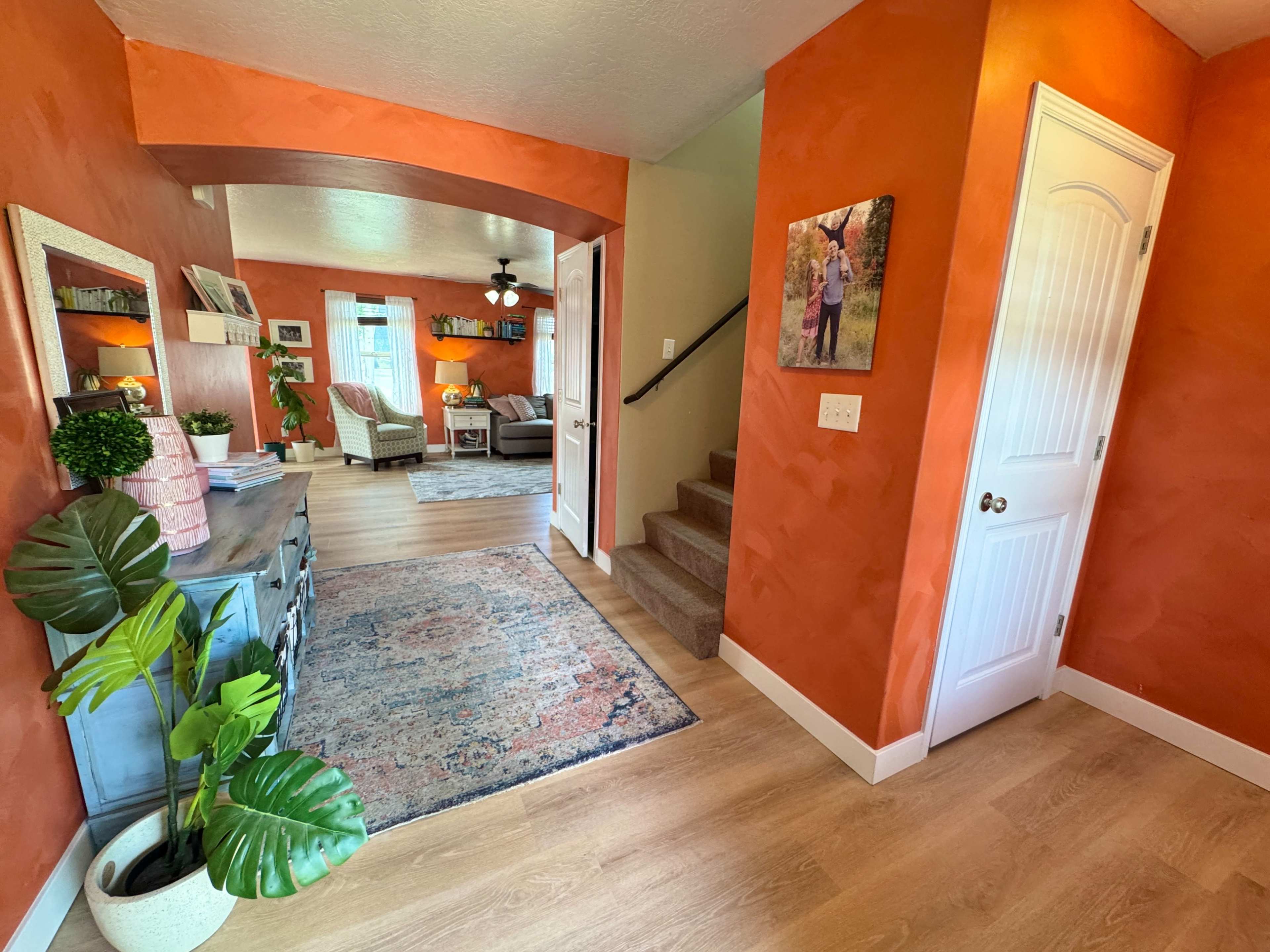 The image shows a warmly decorated entryway with orange walls, a staircase leading to an upper level, and a living area visible in the background.