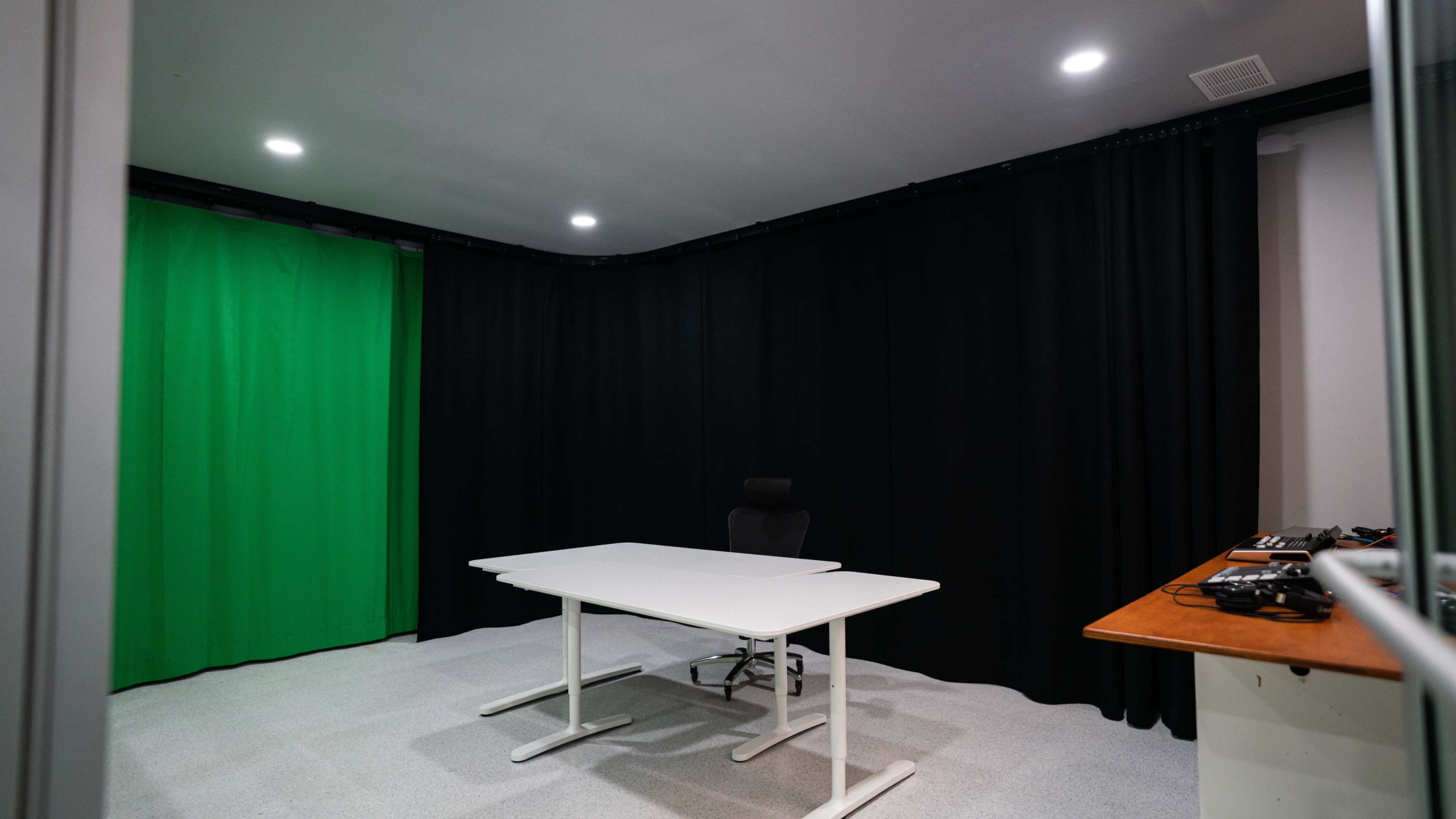 A video production studio with a white table, an office chair, and a green backdrop surrounded by black curtains.