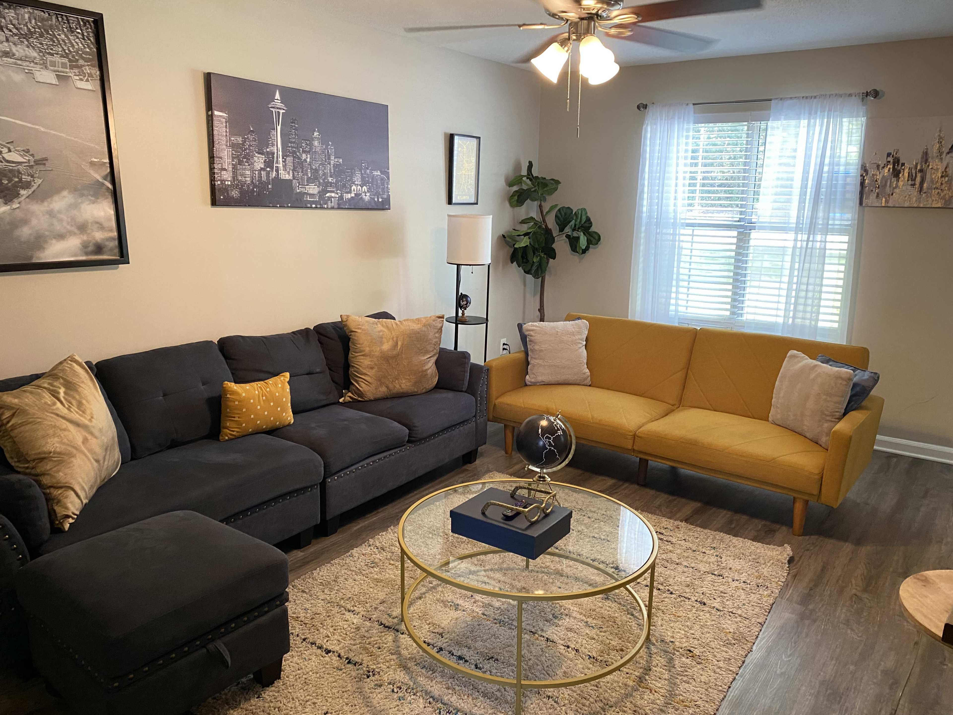 The living room features a dark sectional sofa, a bright yellow couch, and a round glass coffee table, with wall art and a plant enhancing the space.