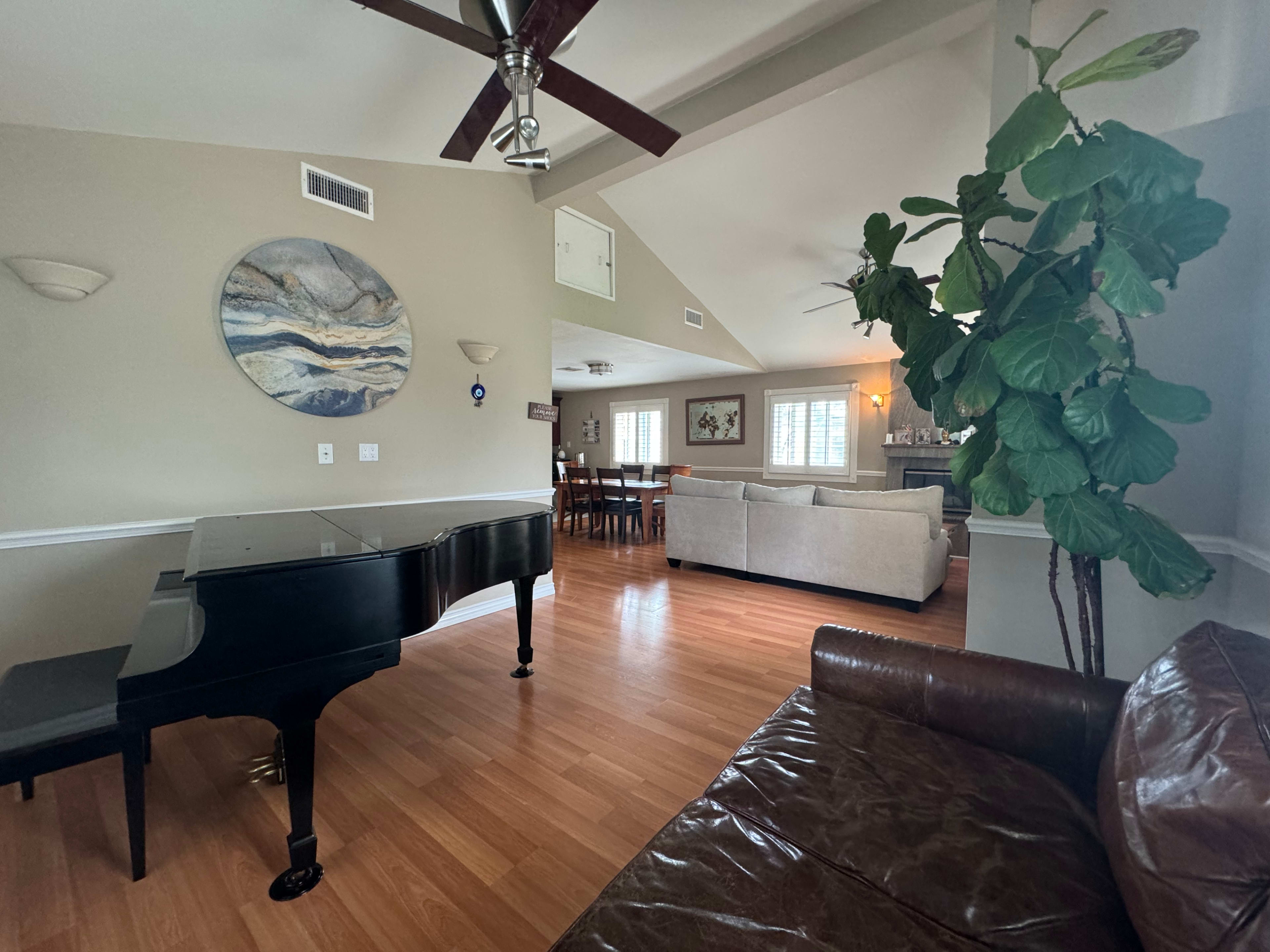 A grand piano sits in the foreground, with a spacious living area featuring a dining table and a couch visible in the background.