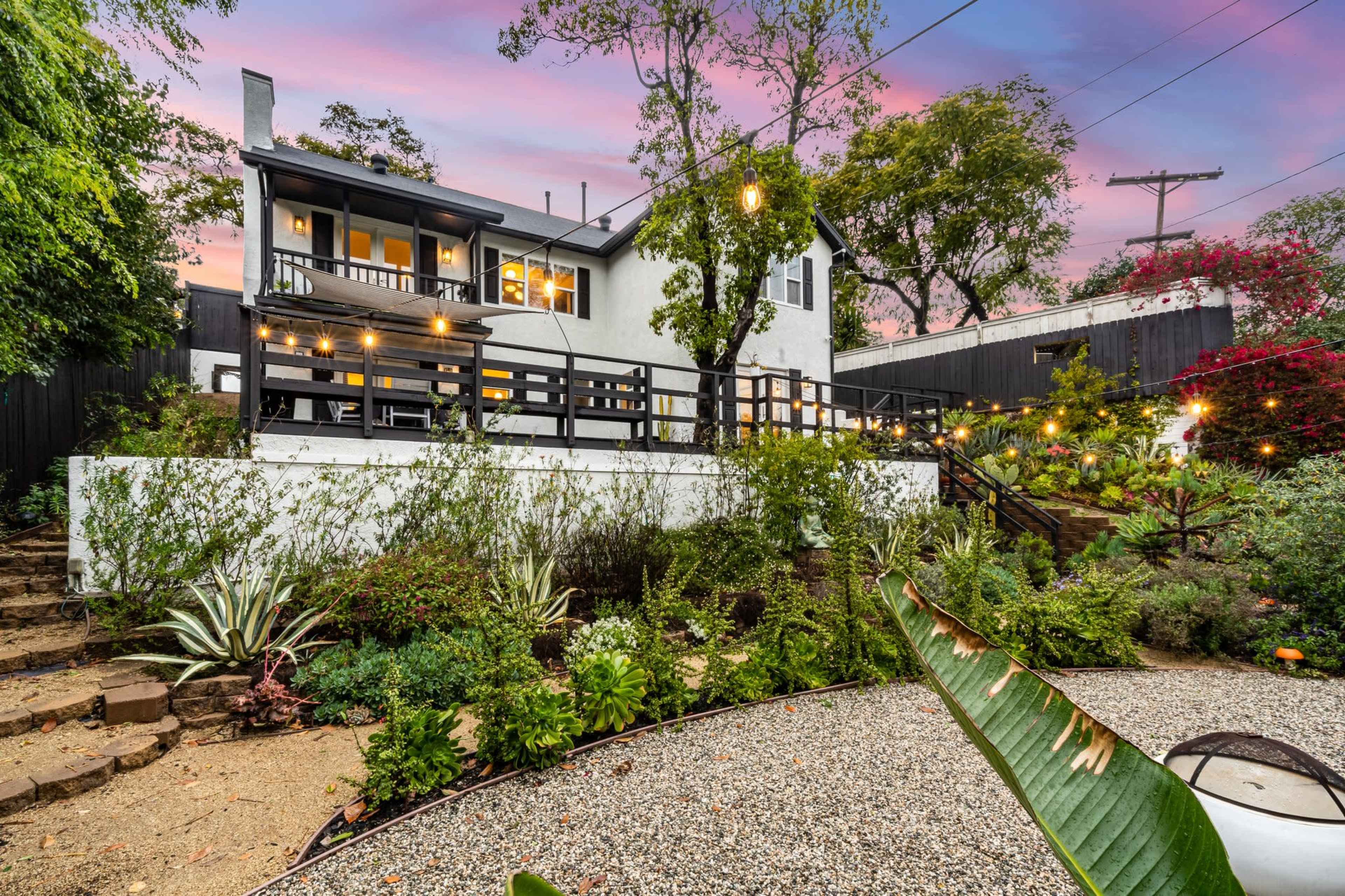A white house with a black deck and string lights overlooks a landscaped garden with various plants and a gravel path.