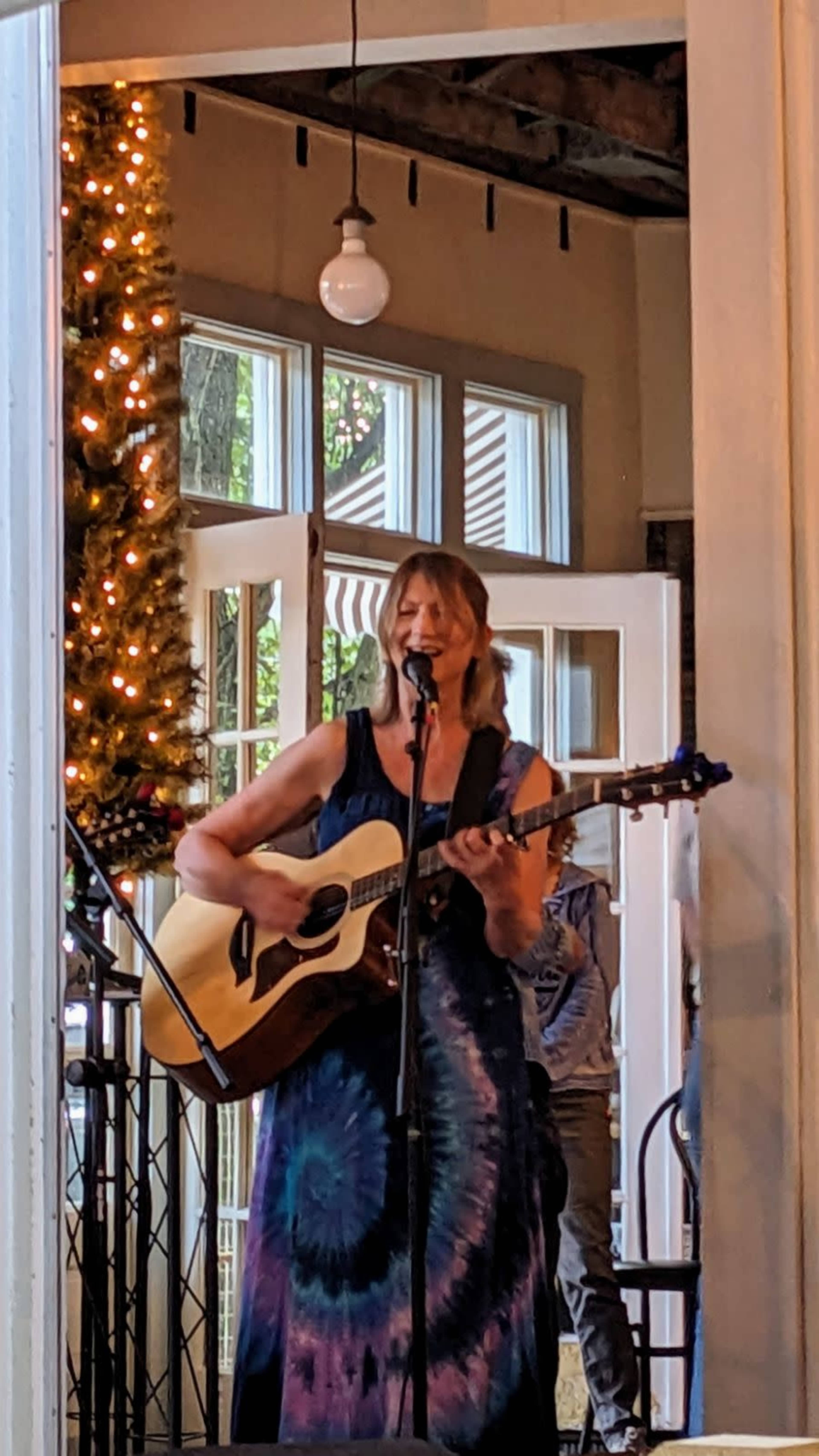 A woman plays an acoustic guitar and sings on stage in a venue decorated with a string of lights.