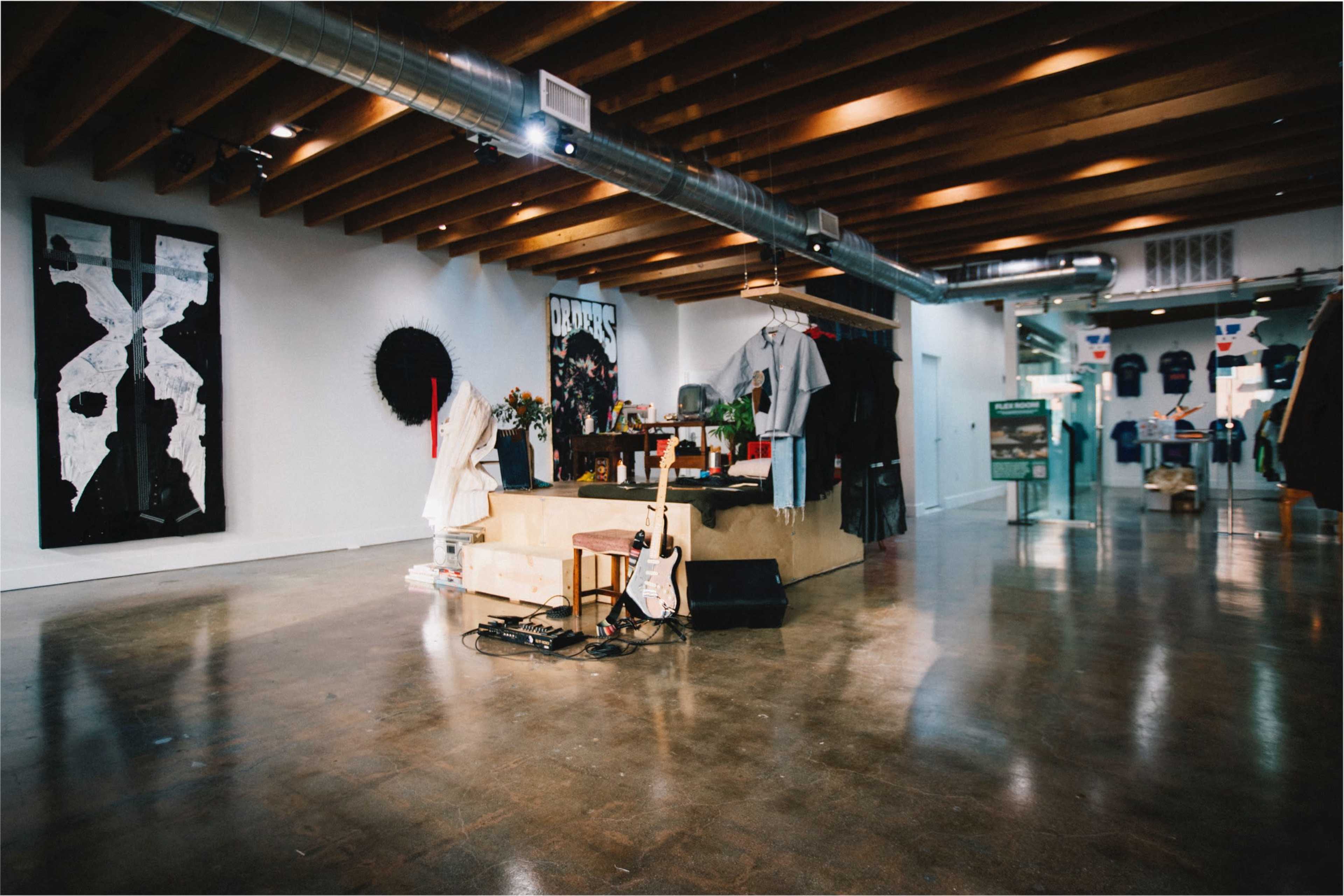 The image shows a modern gallery space featuring a central display of clothing and accessories, with artwork on the walls and polished concrete floors.