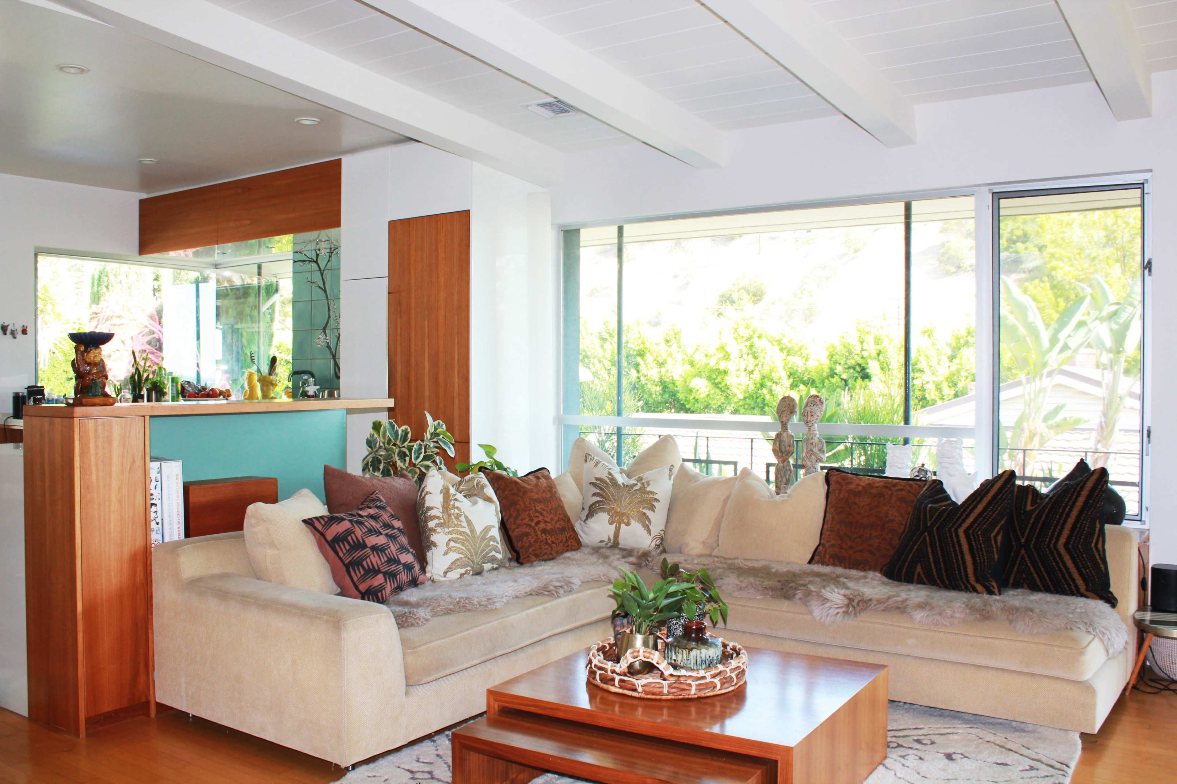A bright living room with large windows, a beige sectional sofa adorned with patterned cushions, and a wooden coffee table.