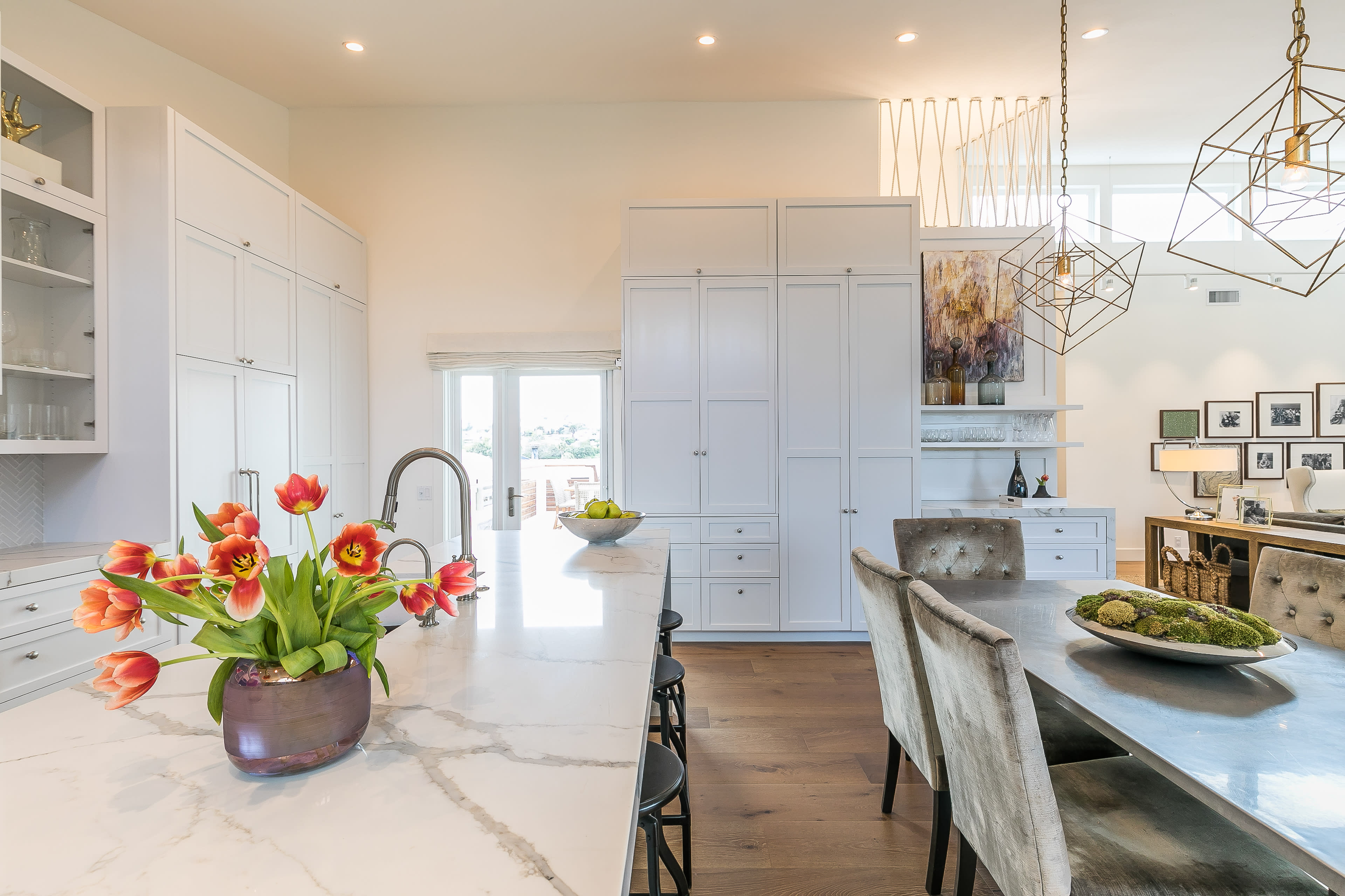 A modern kitchen features white cabinetry, a large marble countertop, and a dining table with a bouquet of tulips.
