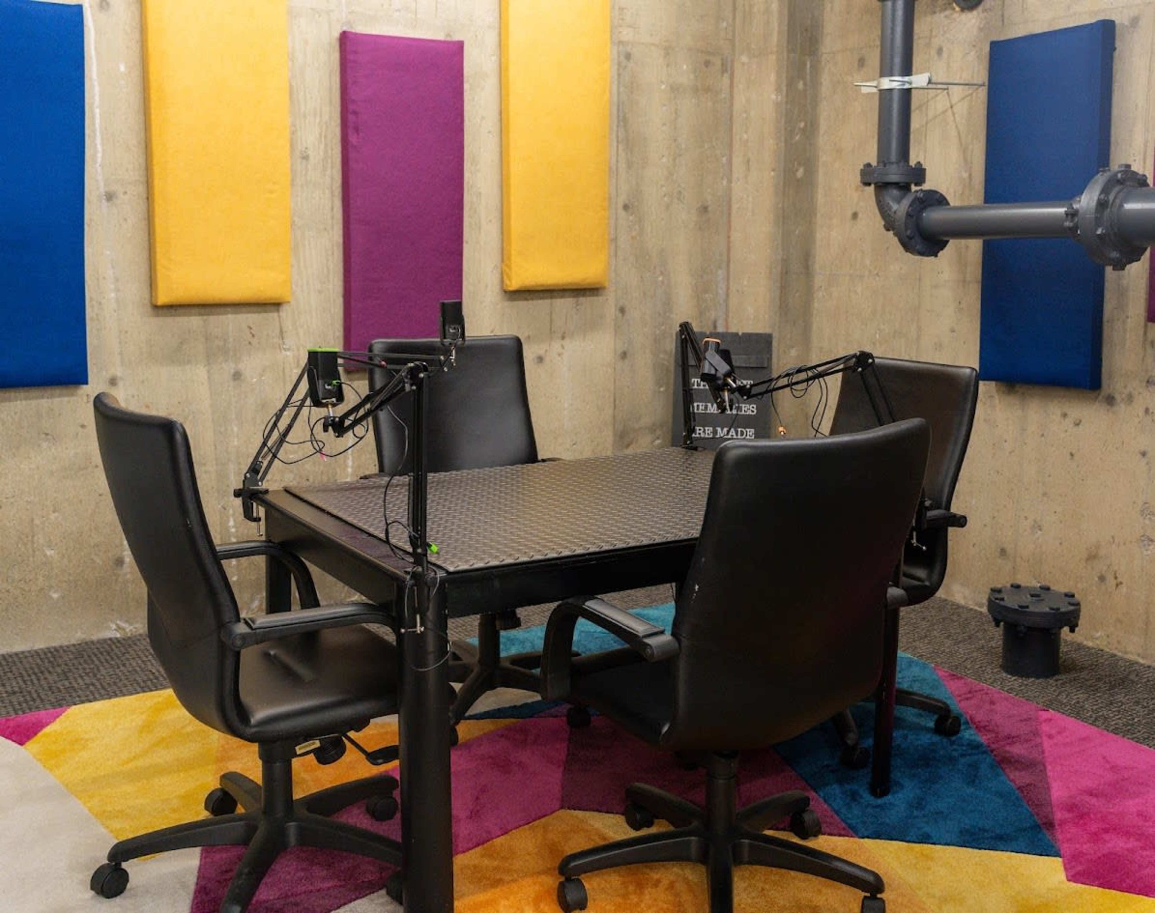 A podcast recording studio features a square table surrounded by four black chairs, with colorful acoustic panels on the walls and a vibrant, patterned carpet.