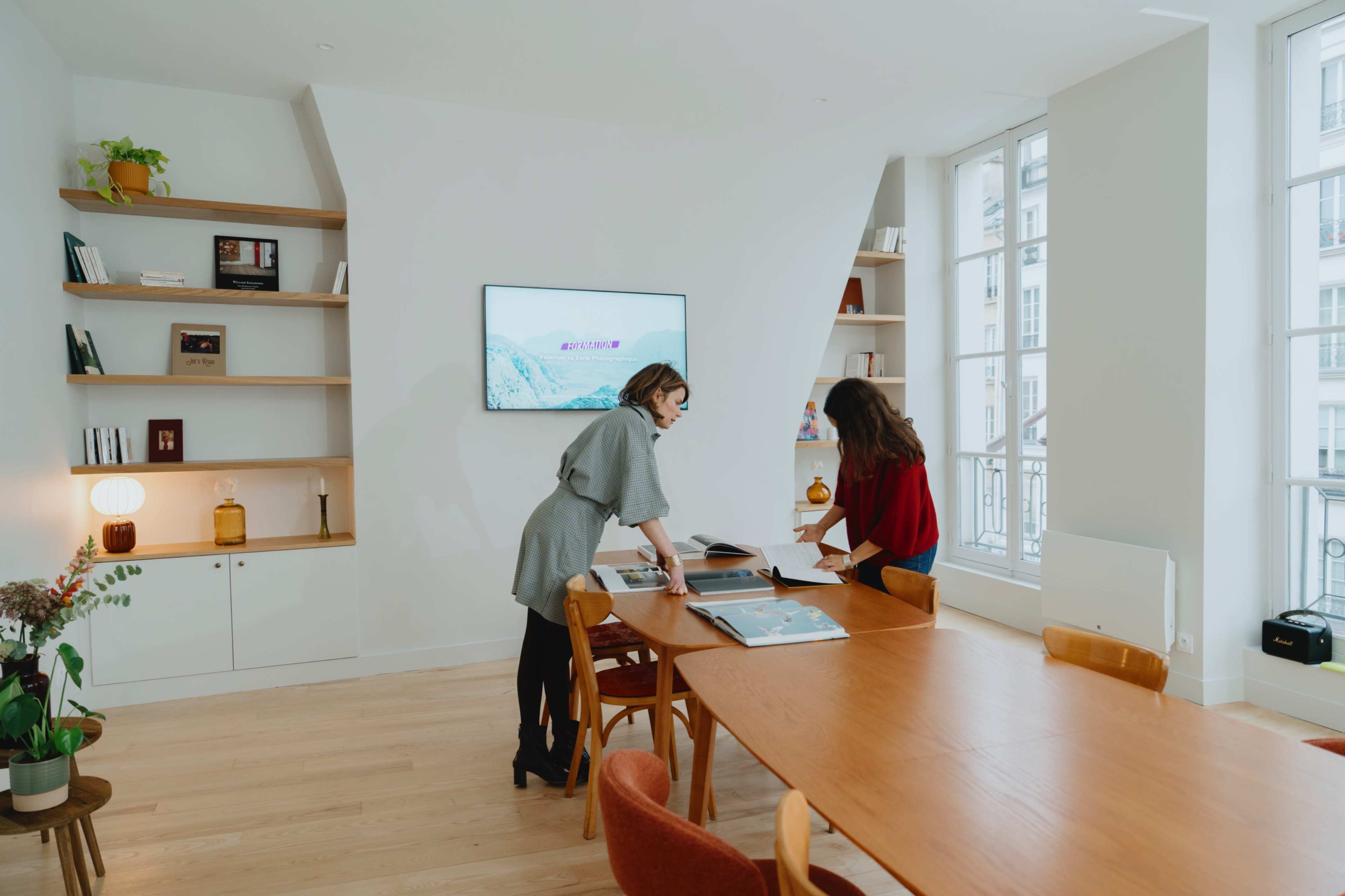 Two women are reviewing documents at a wooden table in a well-lit, modern office with shelves displaying books and decorative items.