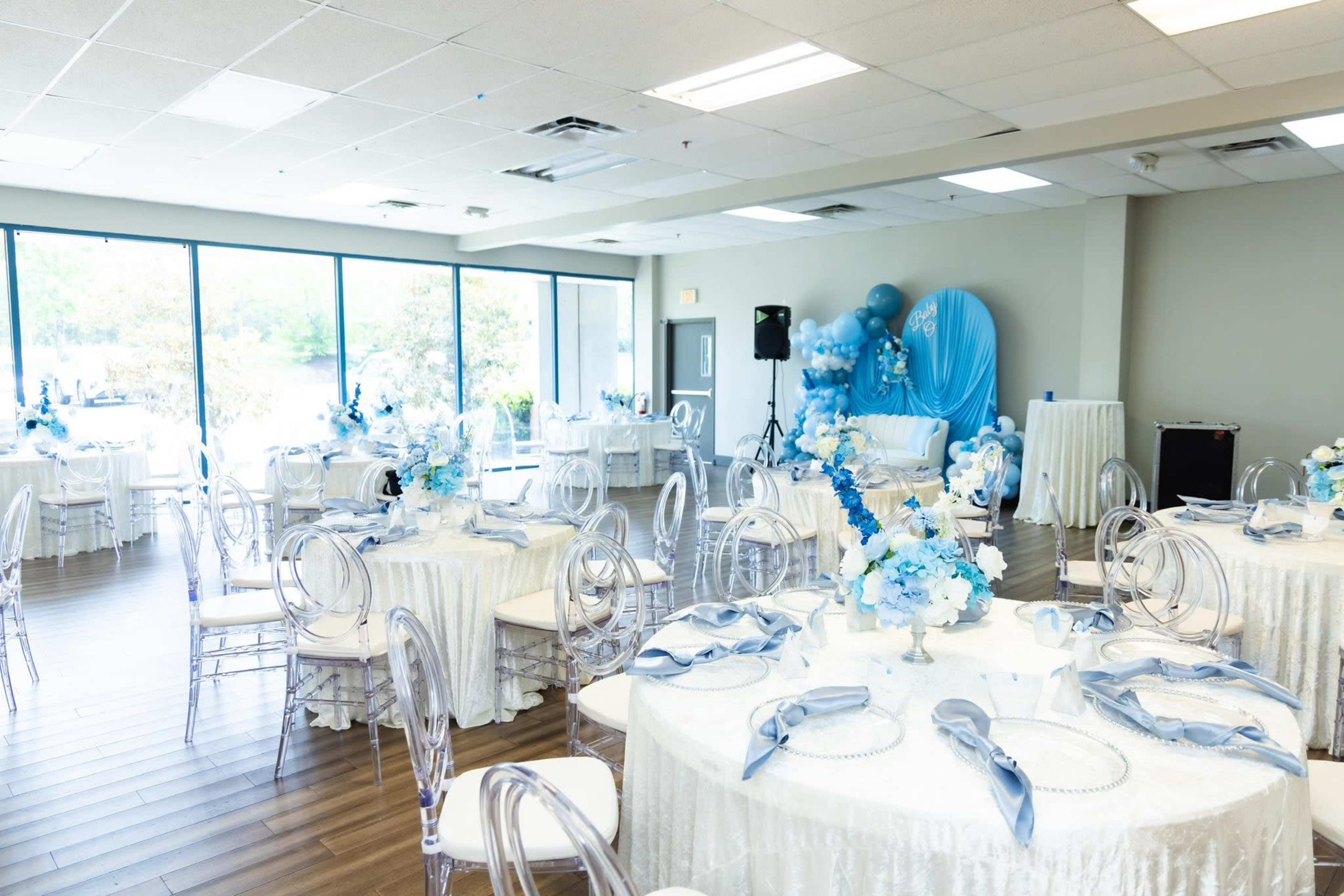 The image shows a decorated banquet hall with round tables covered in white tablecloths, each featuring floral centerpieces in shades of blue.