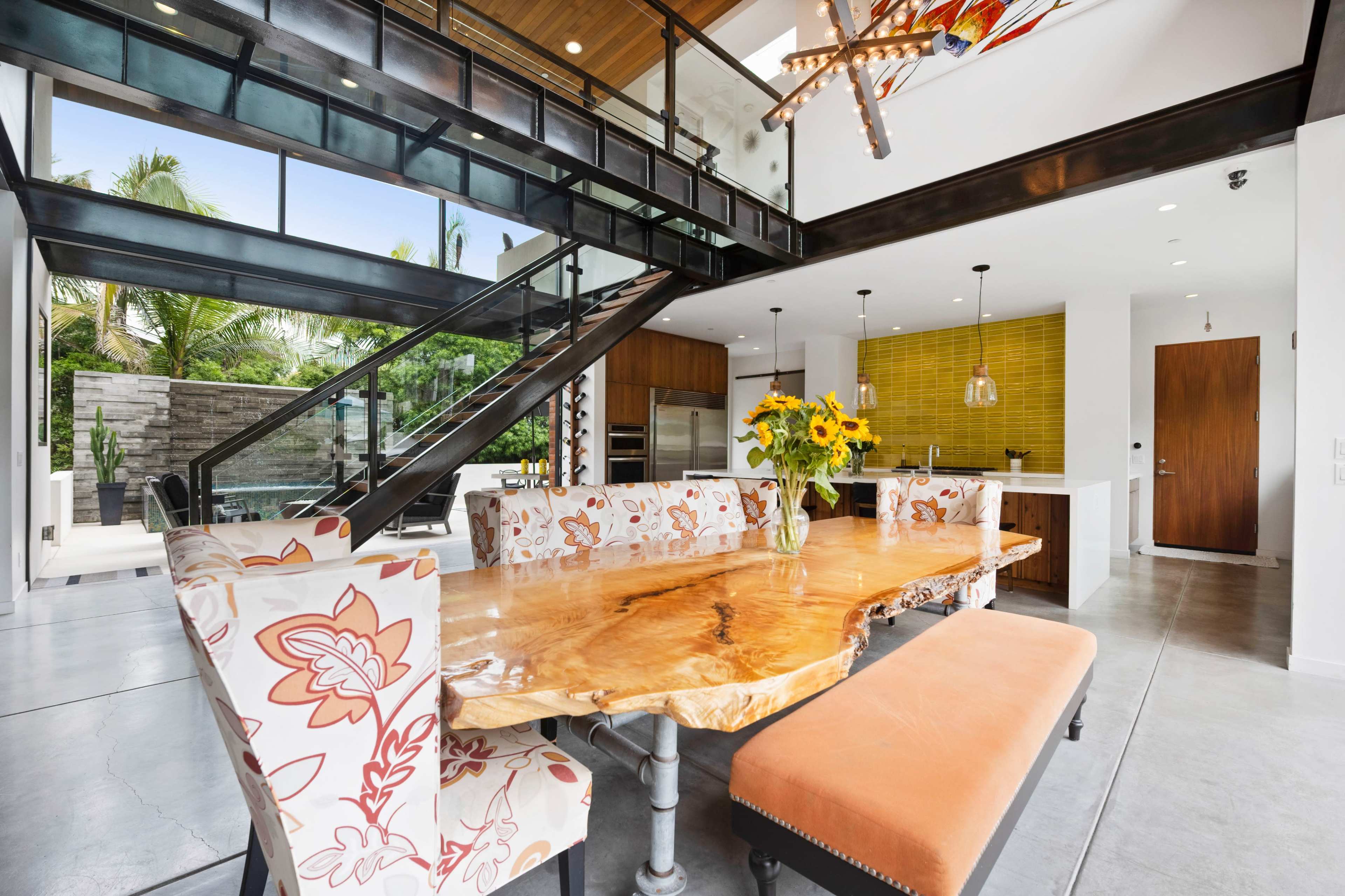 The image features a modern kitchen and dining area with a large wooden table surrounded by floral-patterned chairs, a sleek staircase, and a view of an outdoor space through large windows.