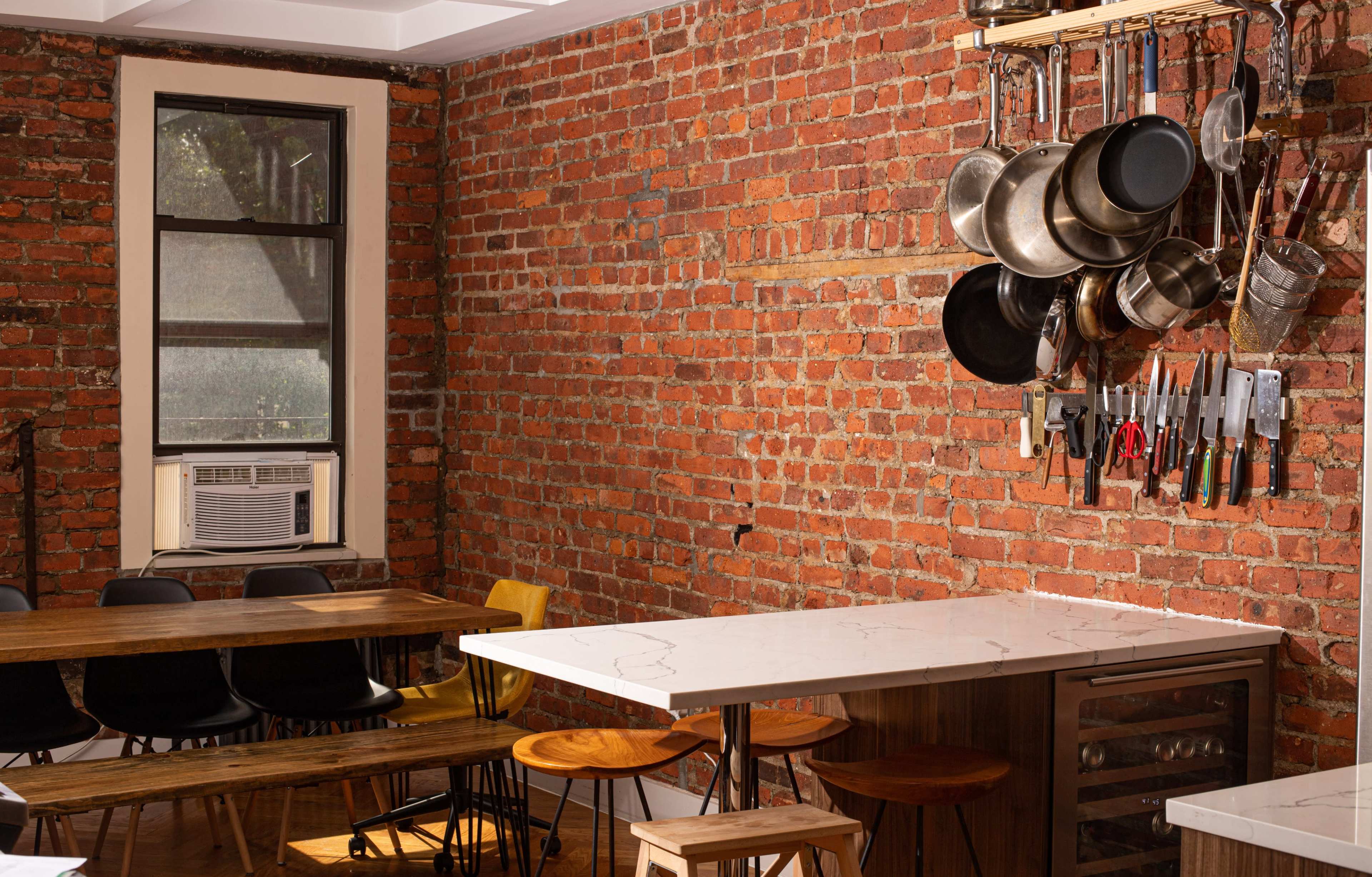A kitchen with exposed brick walls, a countertop, wooden tables, and hanging pots and utensils.