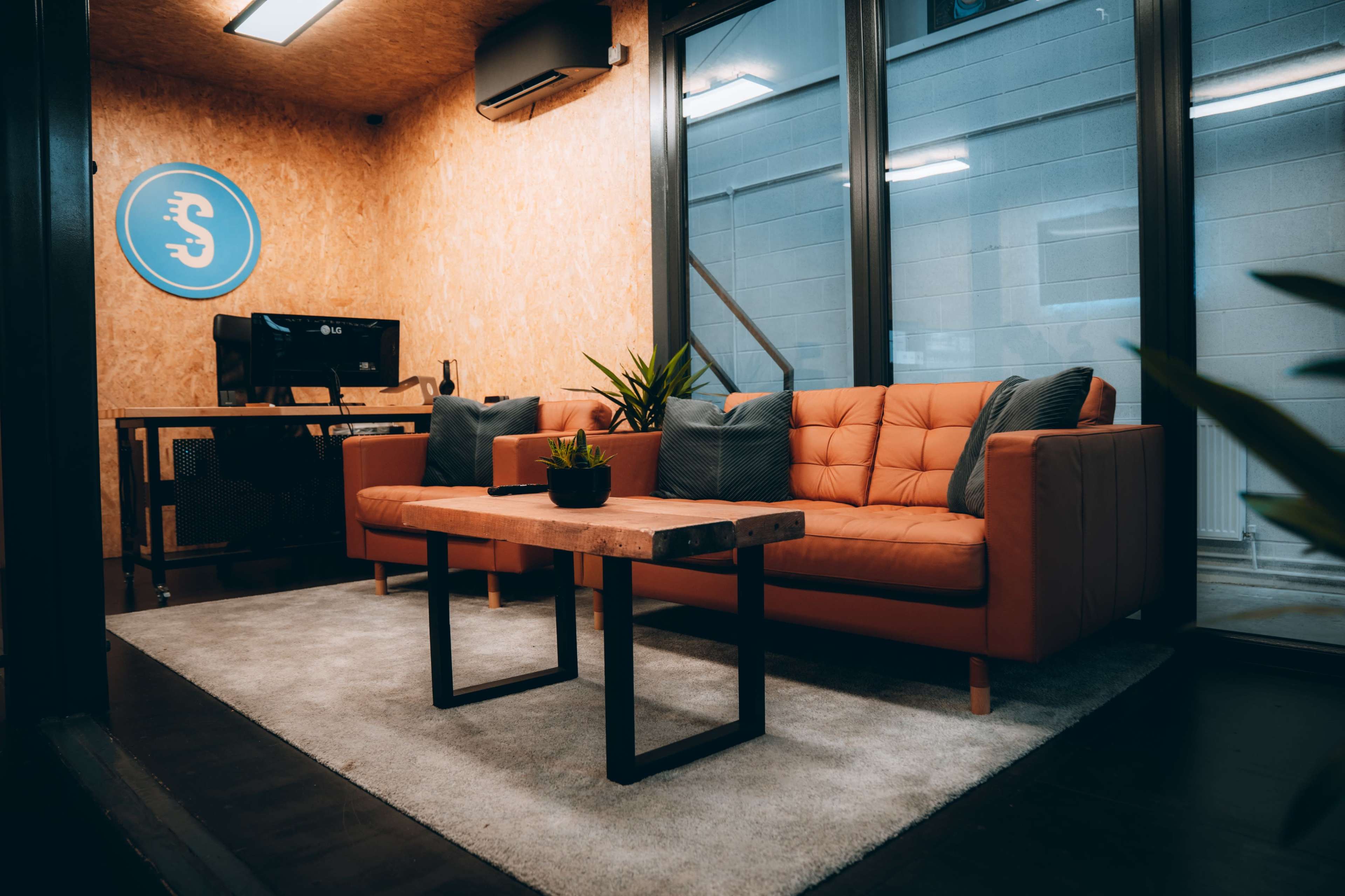 A modern lounge area featuring an orange couch, a wooden coffee table, a small plant, and a desk with a computer, all situated within a room with large windows.