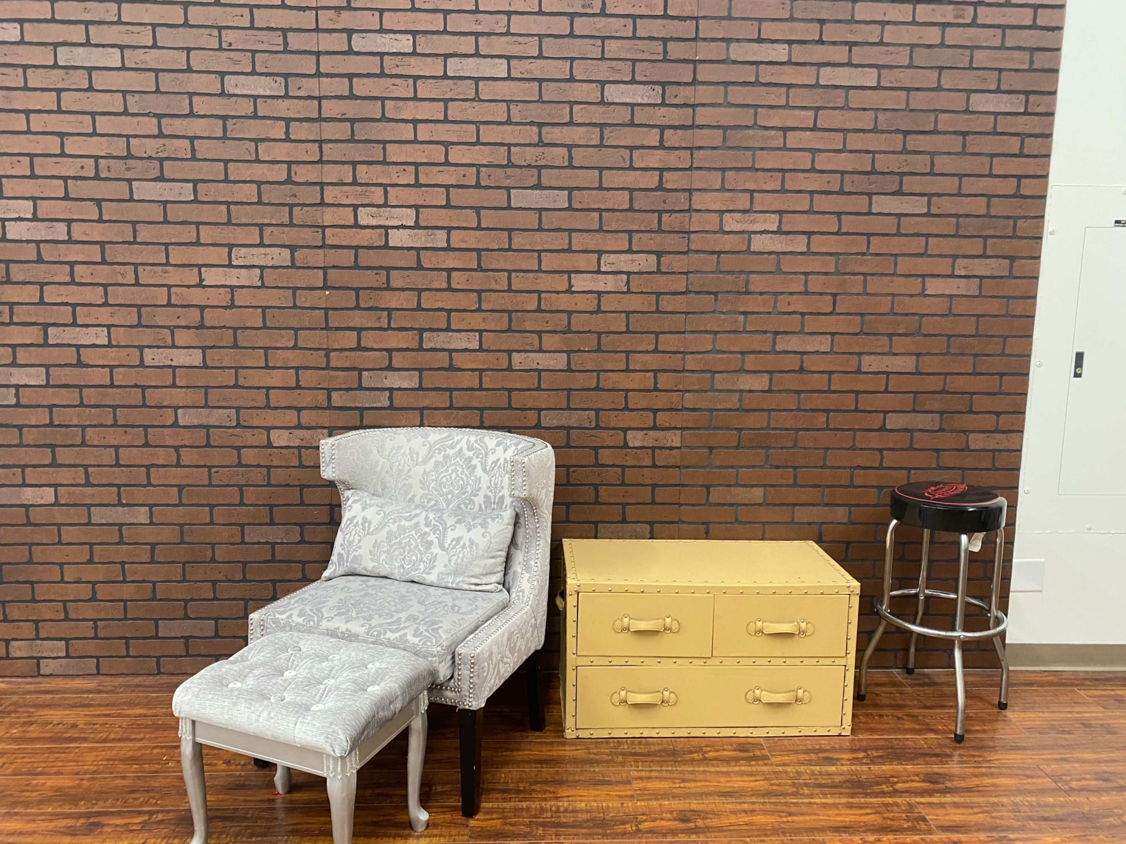 A patterned armchair with an ottoman is positioned next to a yellow storage trunk and a black bar stool against a brick wall.